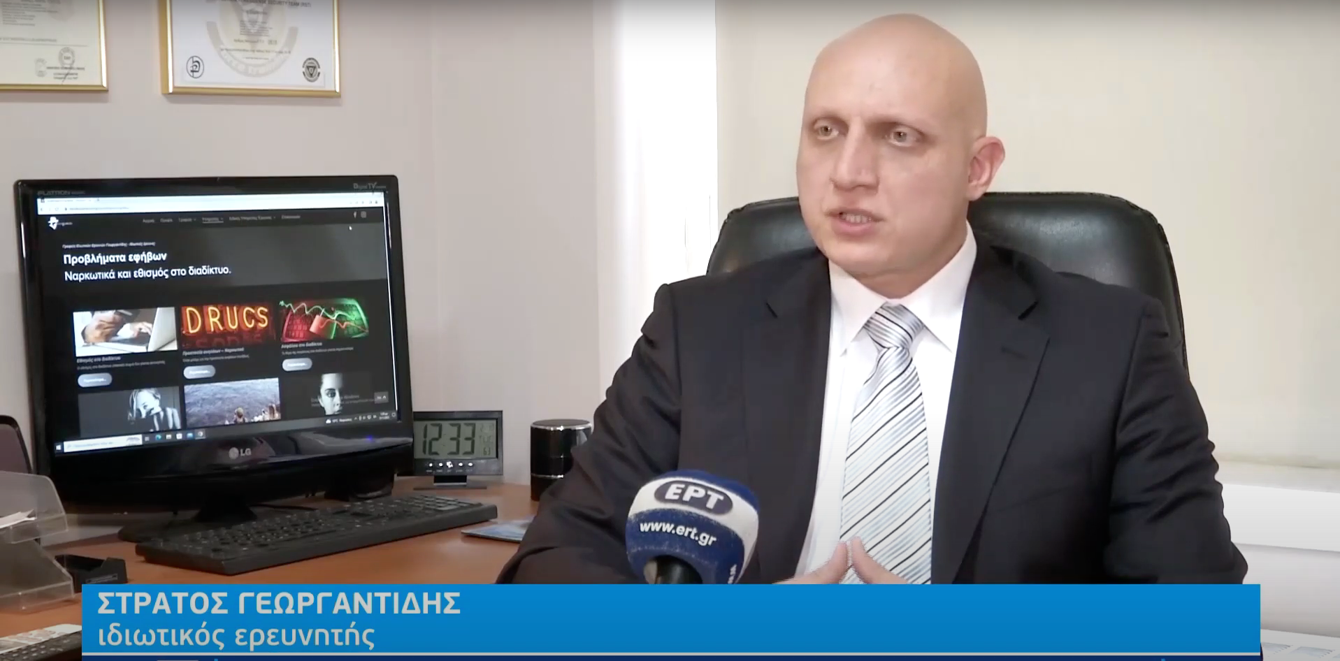 Ένας φαλακρός άντρας με κοστούμι μιλάει σε ένα γραφείο, με μια οθόνη υπολογιστή και ένα μικρόφωνο ορατά.