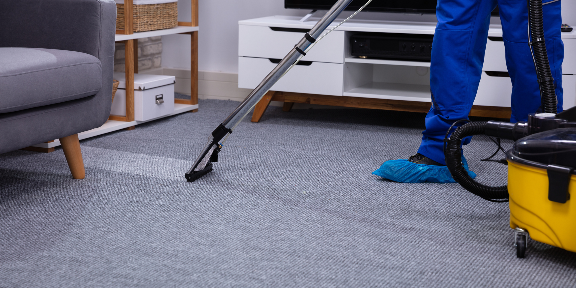 Person vacuuming a grey carpet in a living room, wearing blue coveralls and booties.