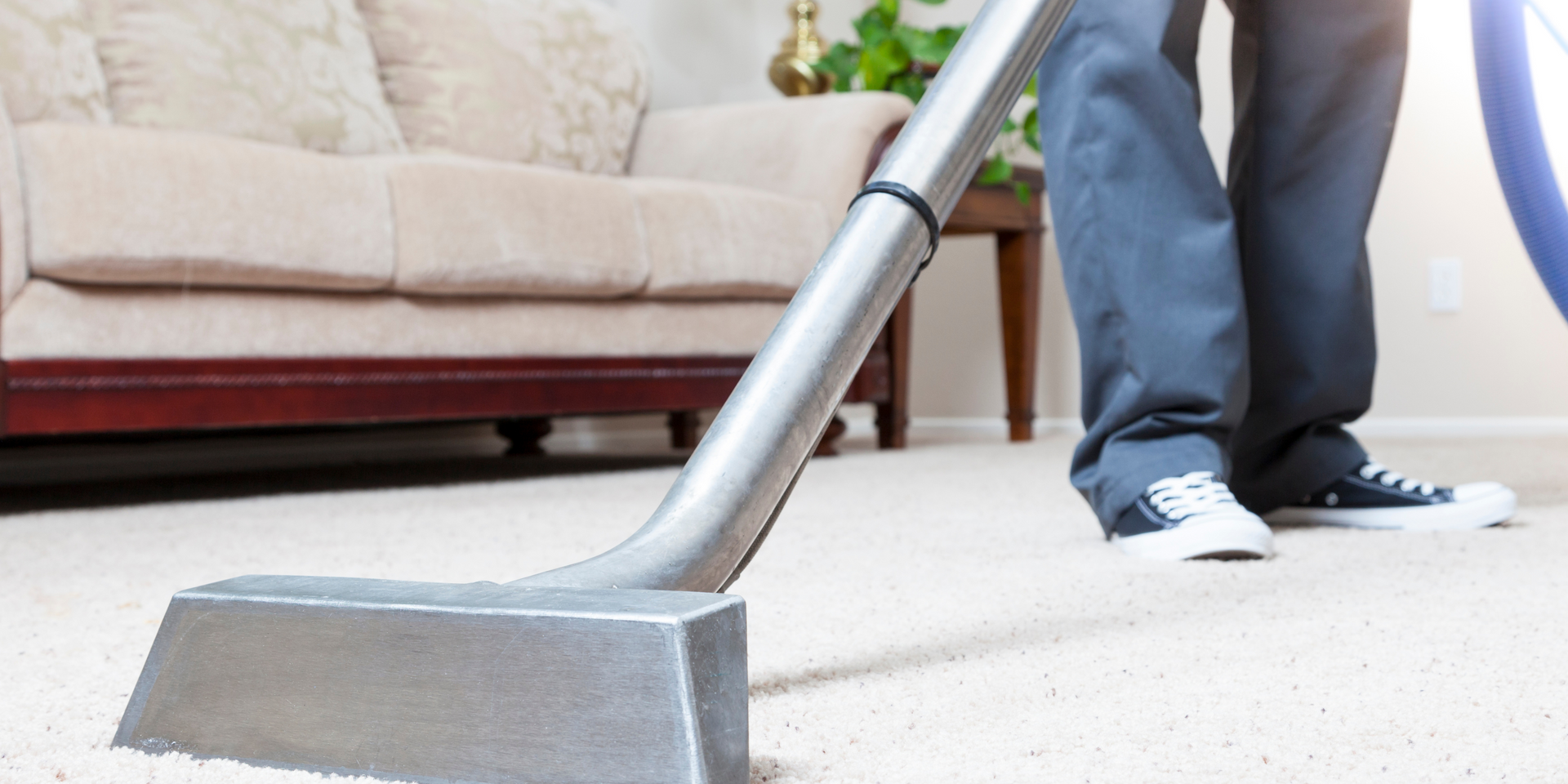A person vacuuming a light-colored carpet in a living room; a sofa is visible in the background.
