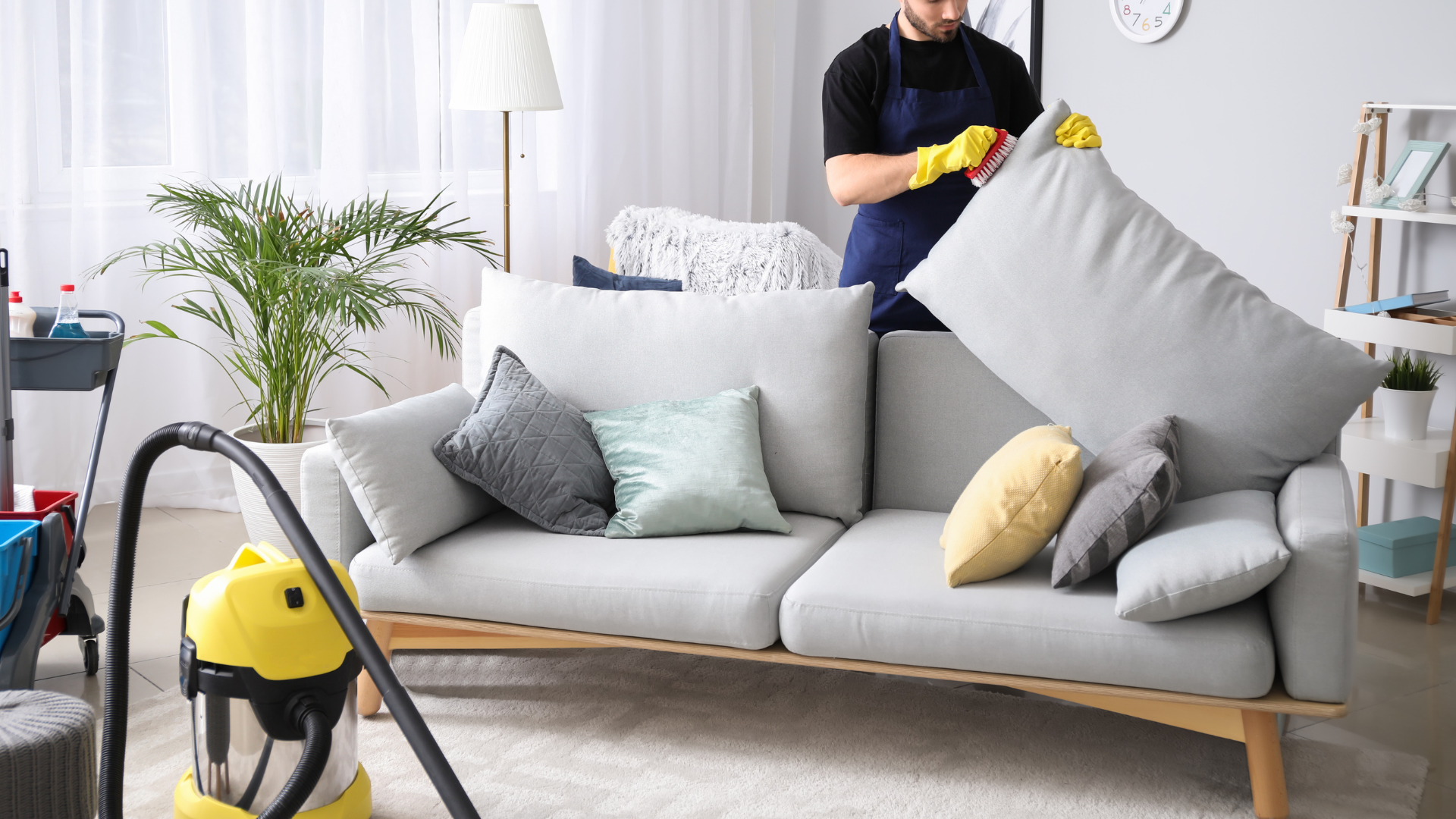 A person in yellow gloves cleans a sofa with pillows, a vacuum cleaner nearby in a living room.