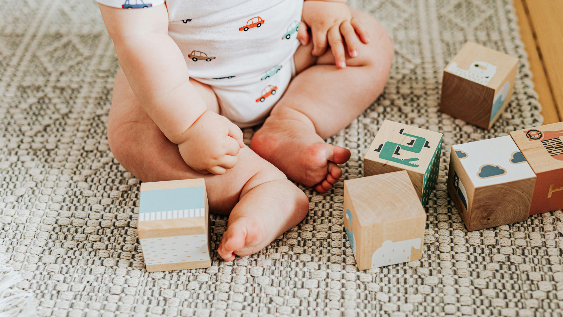 Baby sitting on a patterned rug, playing with wooden blocks.