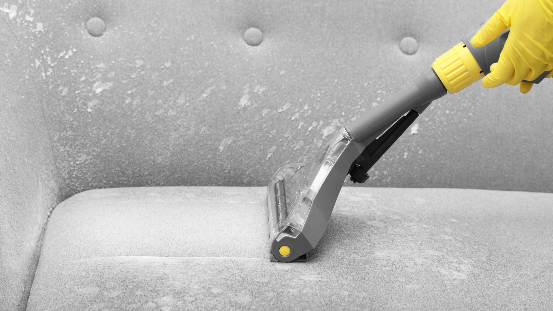 Person vacuuming a gray sofa with a handheld upholstery cleaner, wearing a yellow glove.