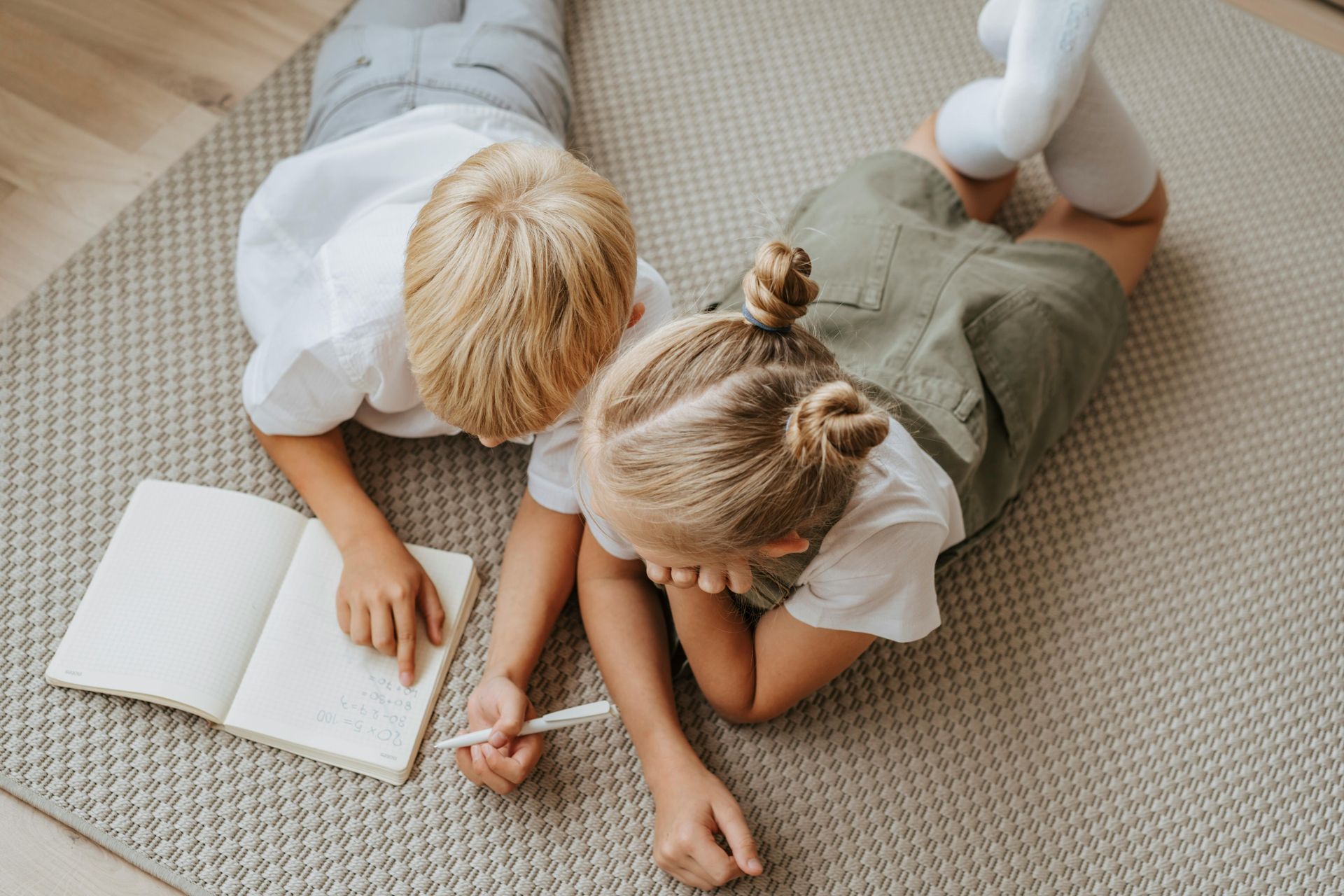 Two children lie on a rug, writing in a notebook together.