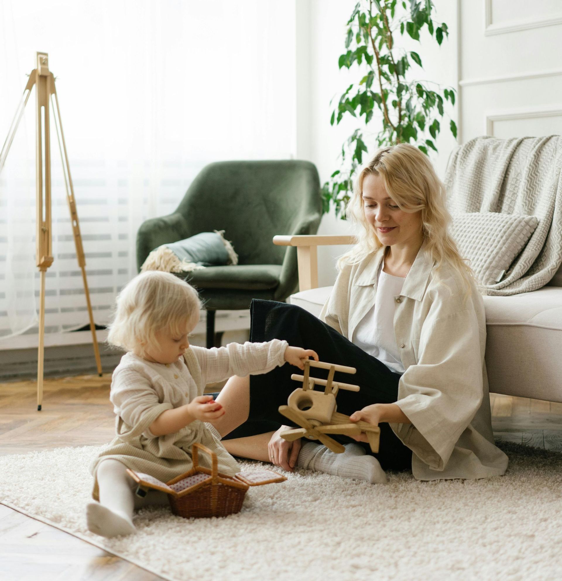 Woman and toddler playing with a wooden toy plane and basket on a rug in a bright room.