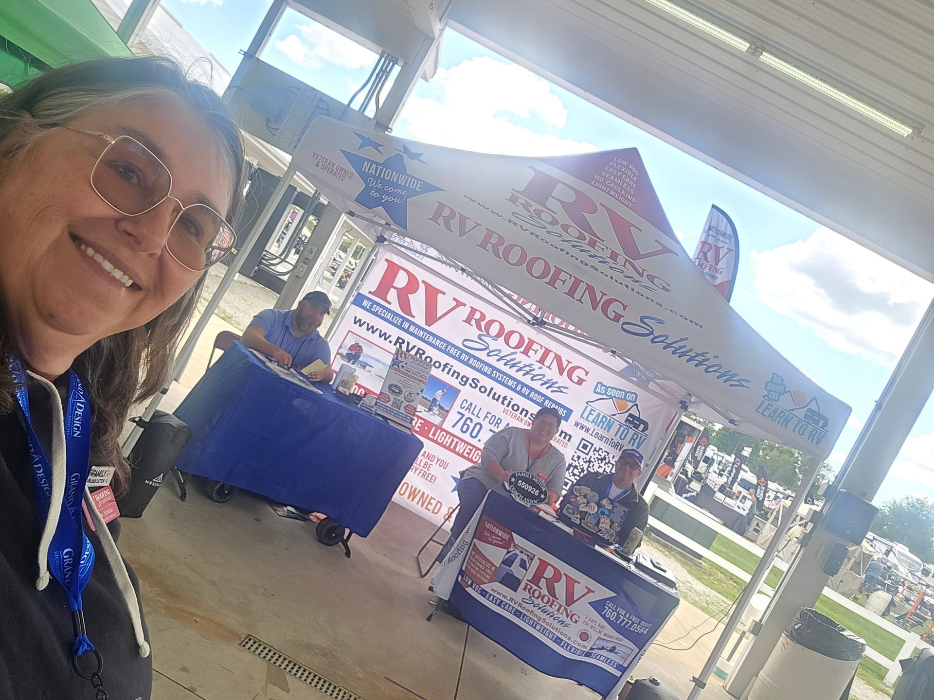 Smiling woman at a booth with RV Roofing Service signage and staff under a white canopy at an outdoor event