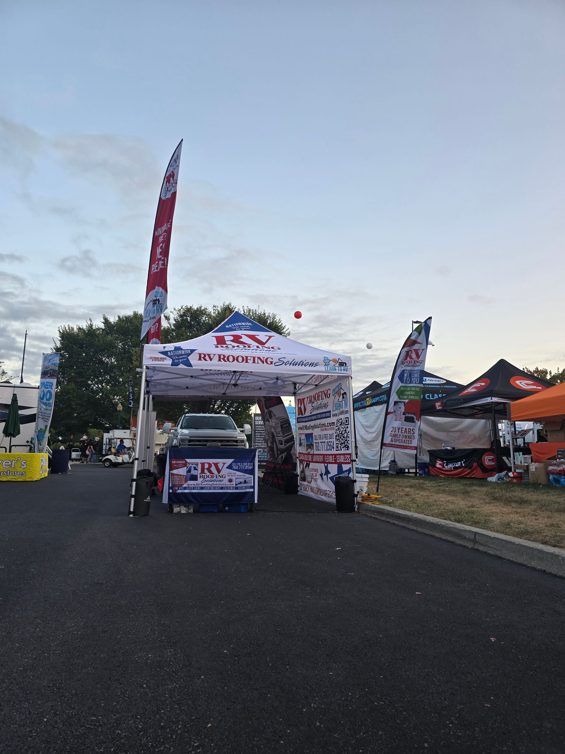Outdoor event booth with a white canopy, banners, and parked vehicle on a paved lot at dusk