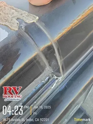 Close-up of a person applying sealant to the corner seam of a dark-colored RV roof. RV Roofing Solutions logo is visible.
