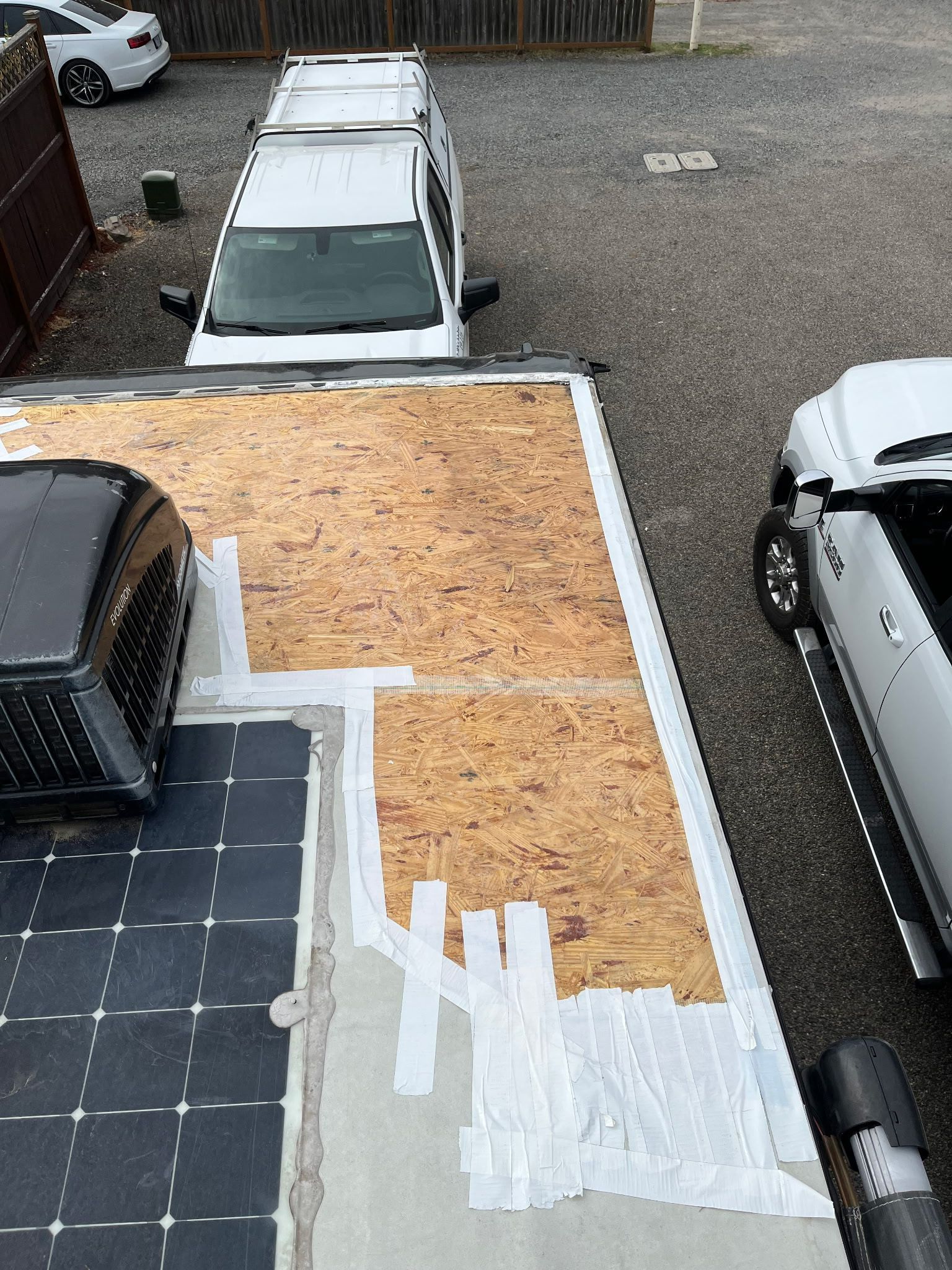 Overhead view of a flat roof with exposed wood, sealant, and a solar panel, with a van and truck parked nearby.