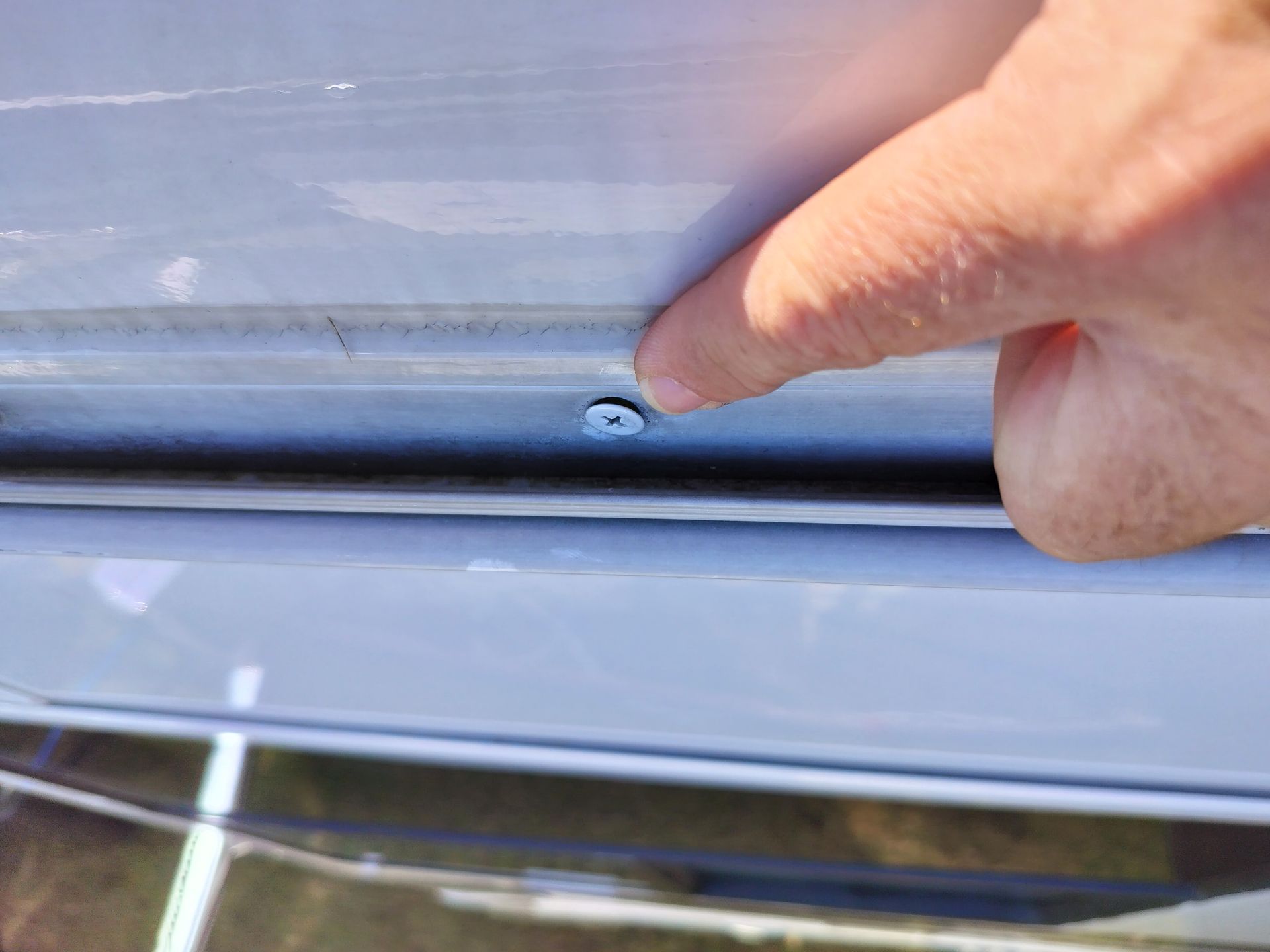 A person's finger pointing at a screw on a light gray metal surface, possibly a car door or trim.