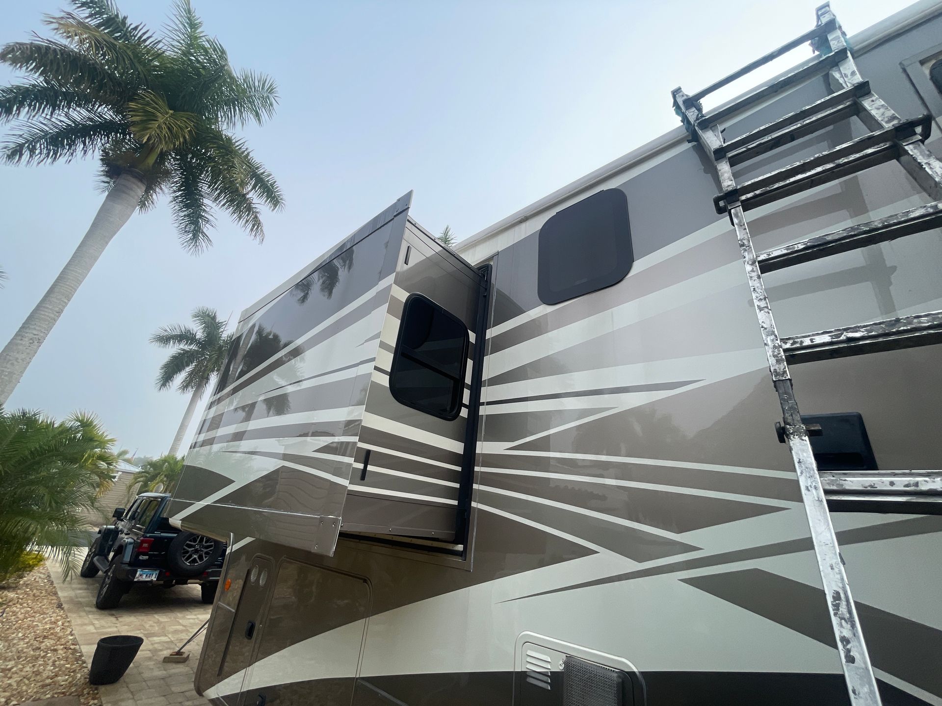 Large gray RV with slide-out and exterior ladder parked beside palm trees on a sunny day