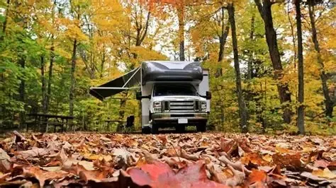 RV parked in a campsite surrounded by trees with fall foliage. Awning extended, leaves on the ground.