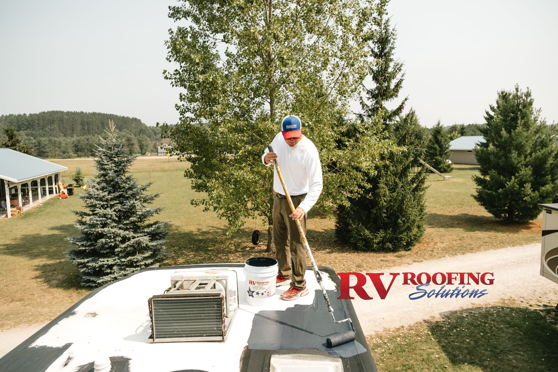 Man on an RV roof, applying white coating with a roller. Outdoors, sunny day.