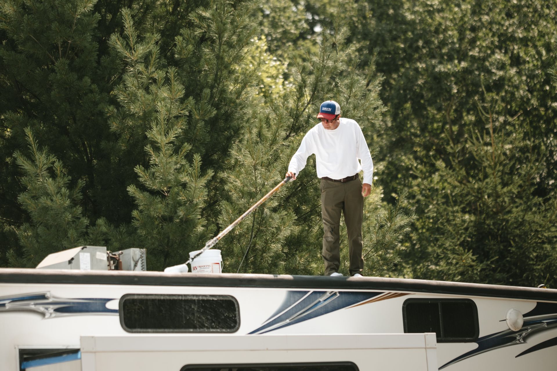 frank on an RV roof, using a tool. Wearing white shirt, blue hat, green pants. Surrounded by trees.
