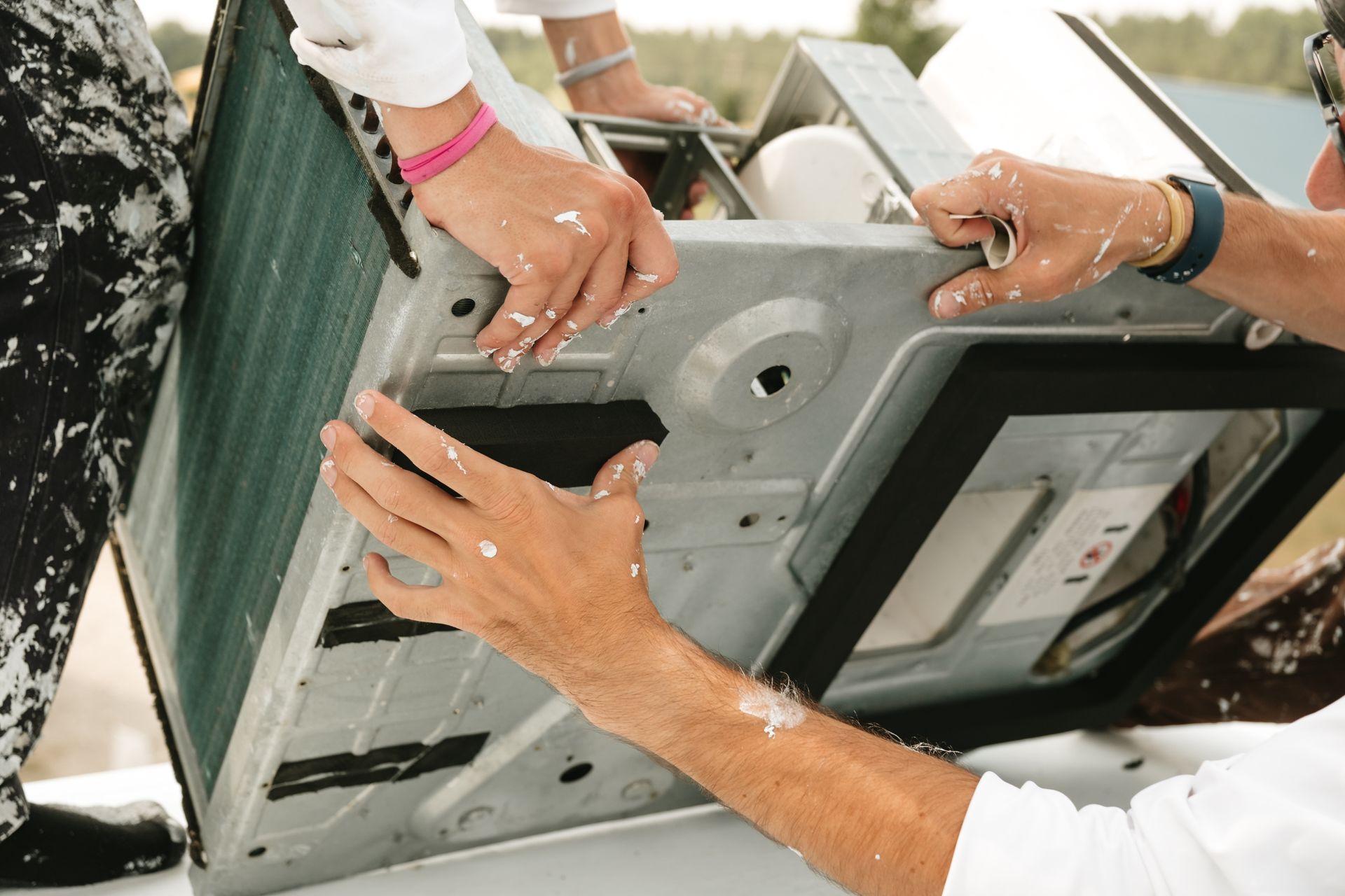 Three people installing an air conditioning unit; hands covered in paint, outdoors.