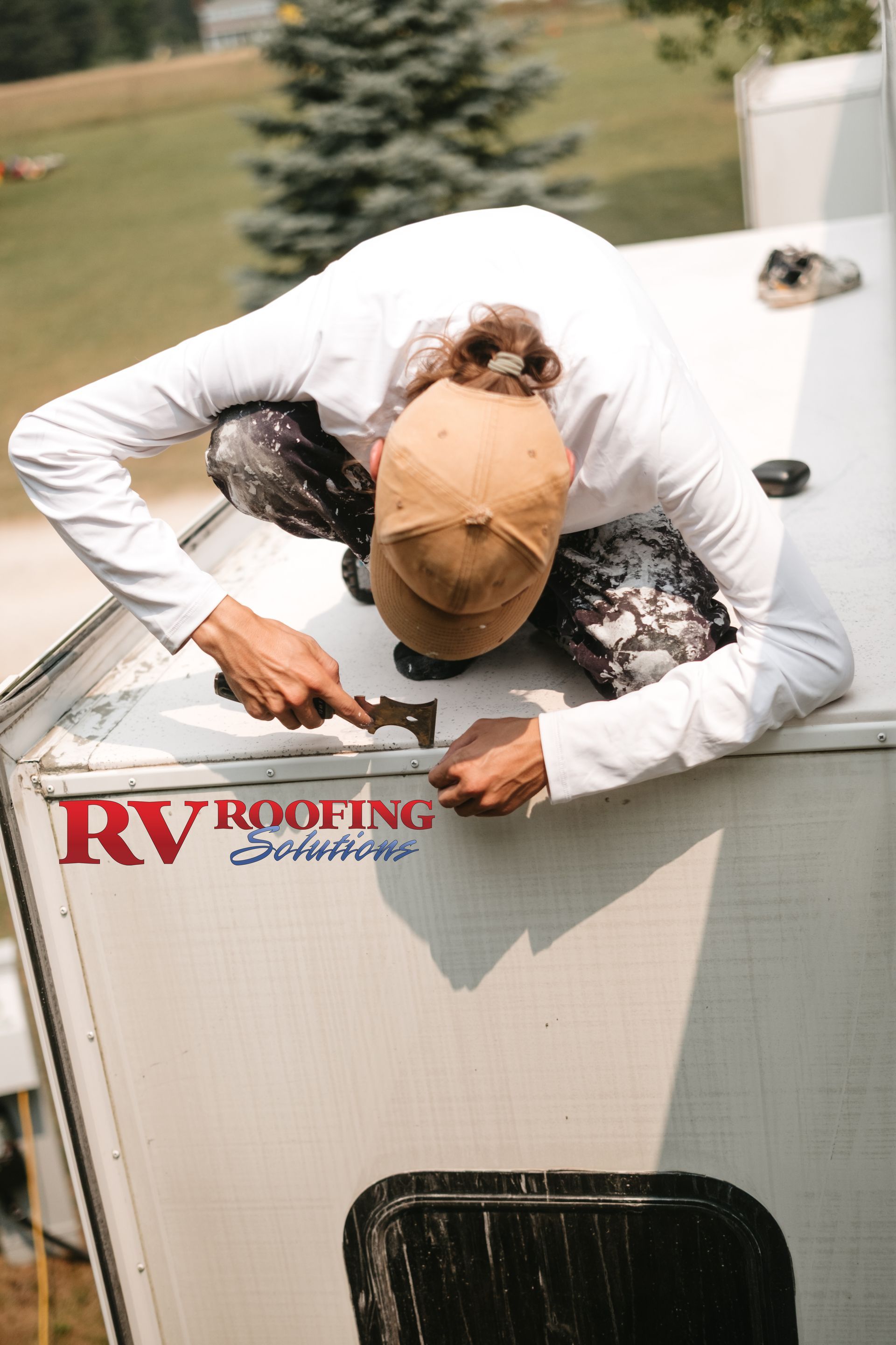 Person on RV roof, repairing, wearing hat, white shirt, outdoors, RV Roofing logo visible.