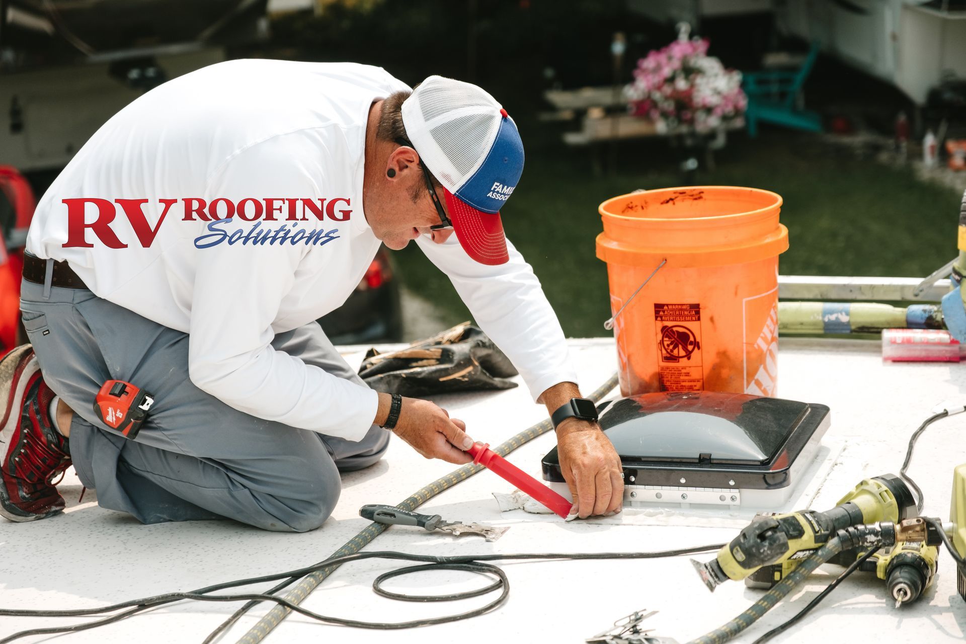 A person repairs an RV roof, kneeling on top. They wear a white hat and are using a tool.