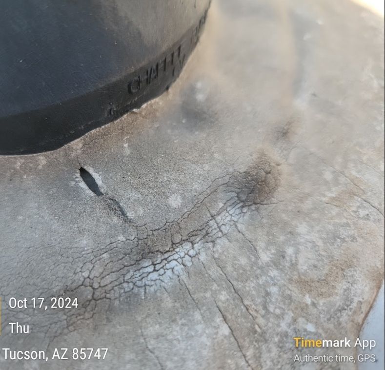 Close-up of a cracked, light-colored roofing material around a black pipe with a slit and a bulge.