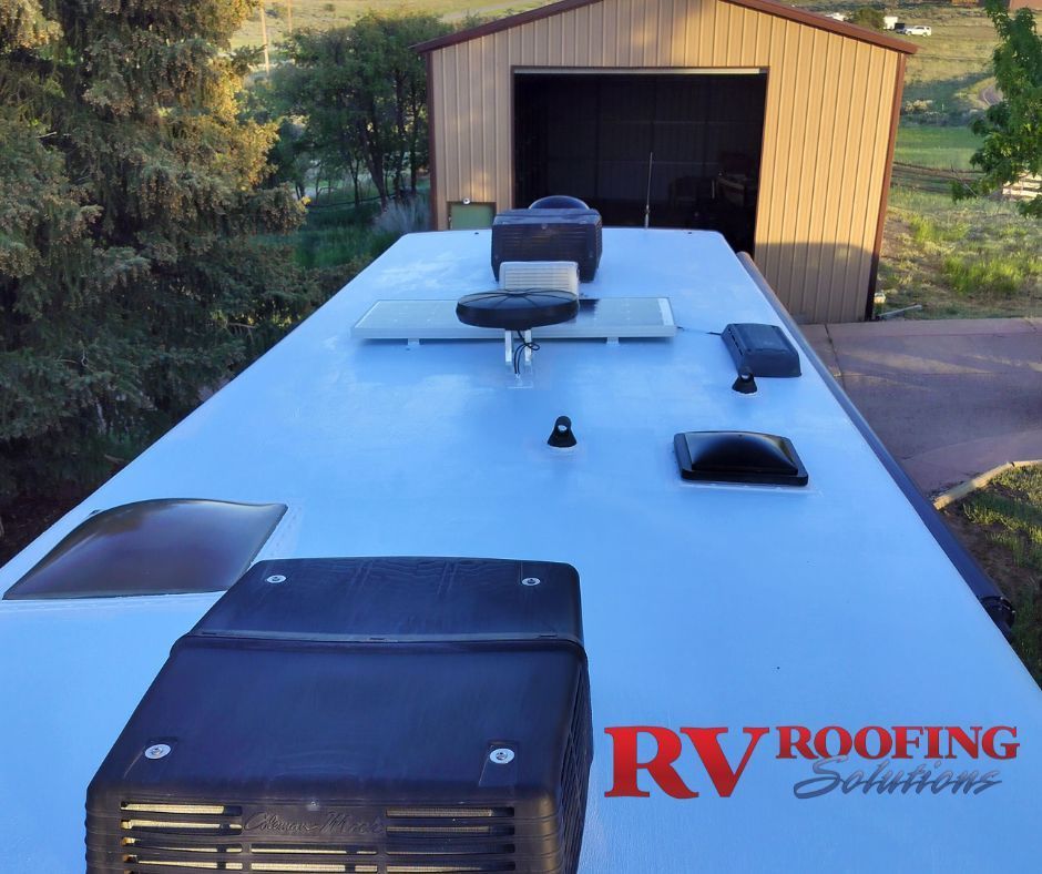 RV roof with black vents and fixtures. Building and trees in the background.