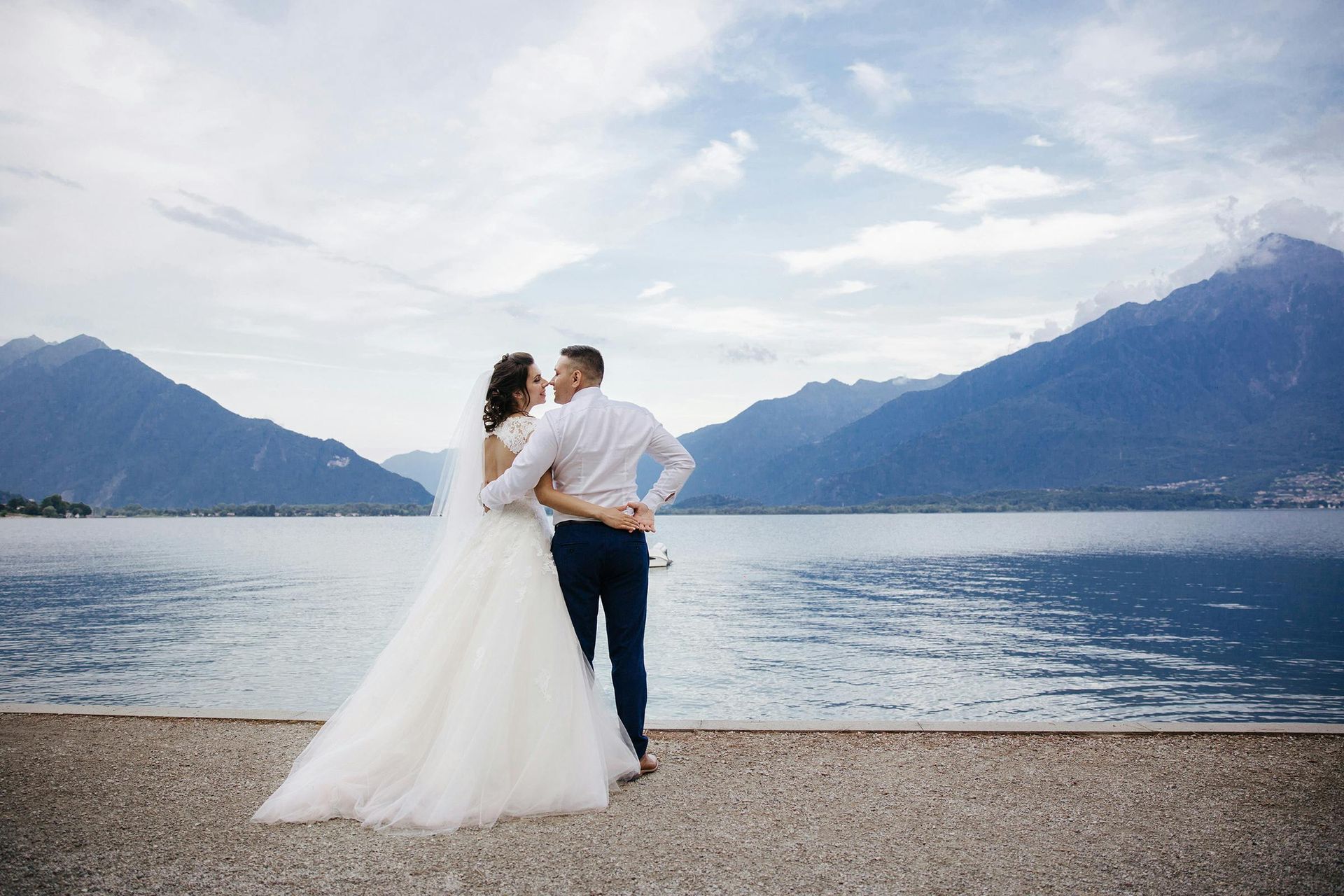 A bride and groom are standing on a beach looking at a lake.