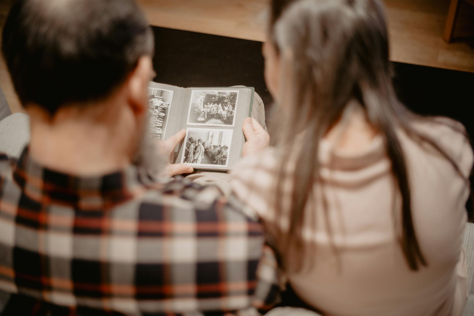 Couple looks at a photo album together, soft focus. Man in plaid, woman in beige. Indoor setting.