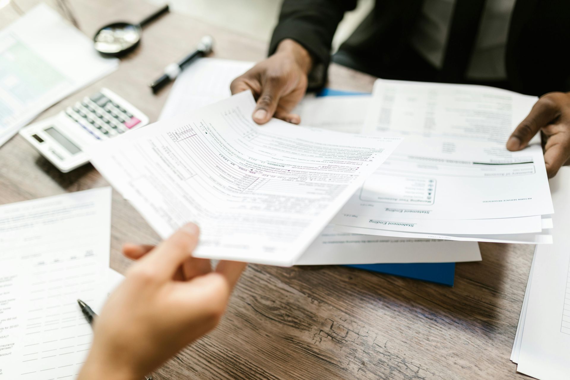 Two people exchanging documents at a desk with a calculator and magnifying glass.