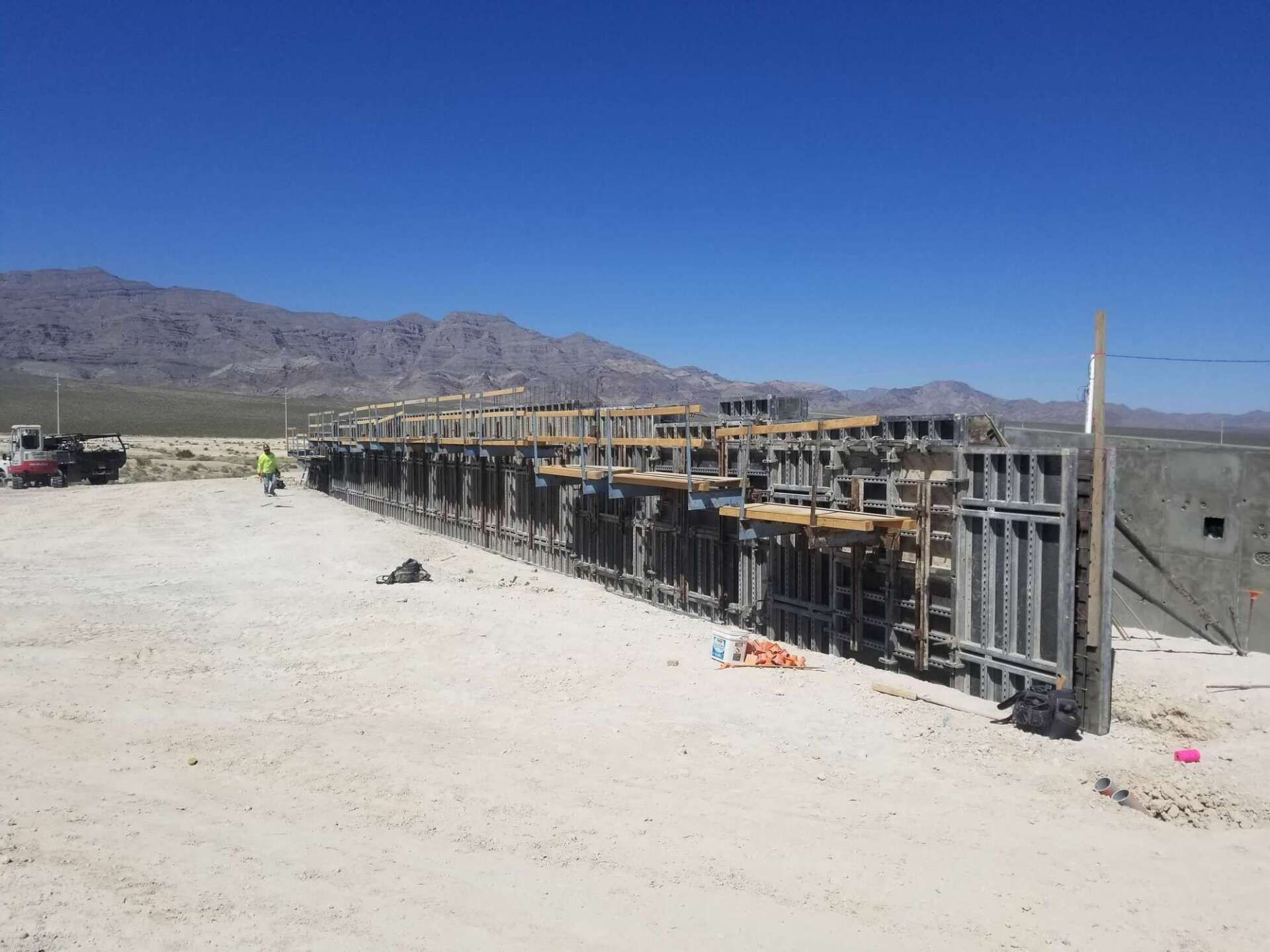 Construction site: Concrete forms and wooden scaffolding set against a mountain backdrop under a blue sky.