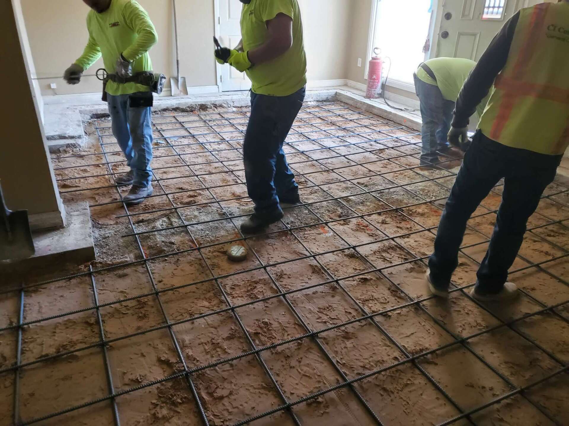 Construction workers installing rebar on a subfloor in a room. Men in neon shirts, safety vests.