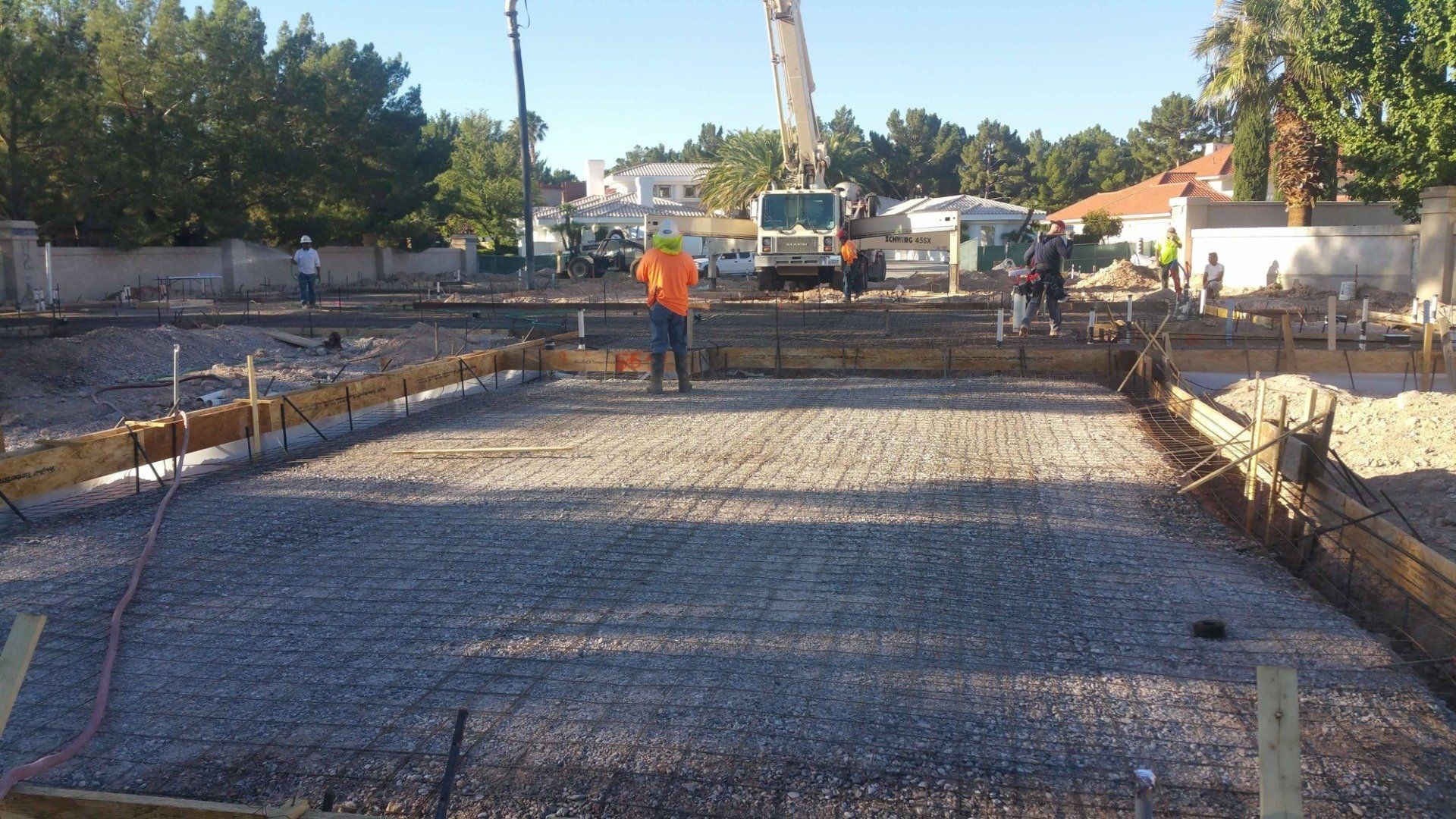 Construction site: Workers pouring concrete foundation. Gravel, forms, truck, and boom visible. Sunny day.