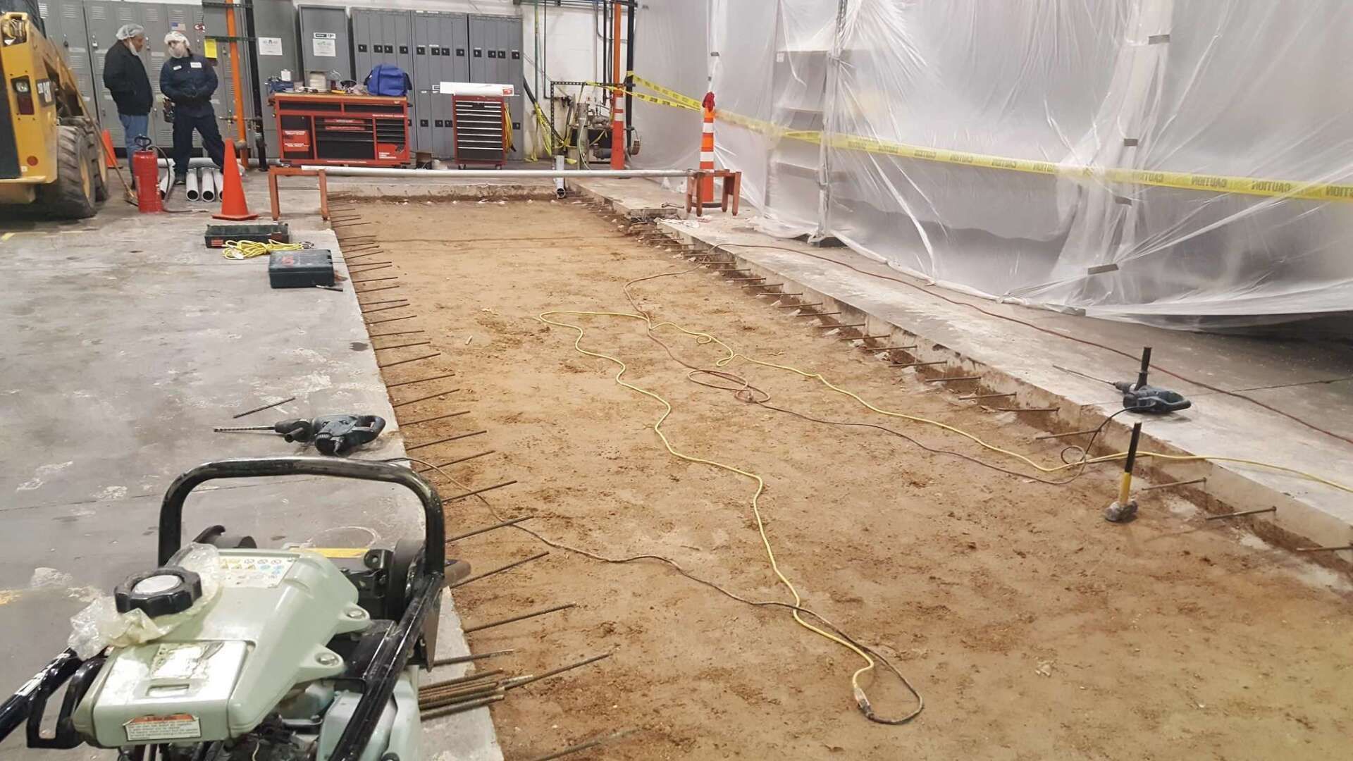 Construction site with exposed ground. Workers observe near equipment; yellow caution tape surrounds the area.
