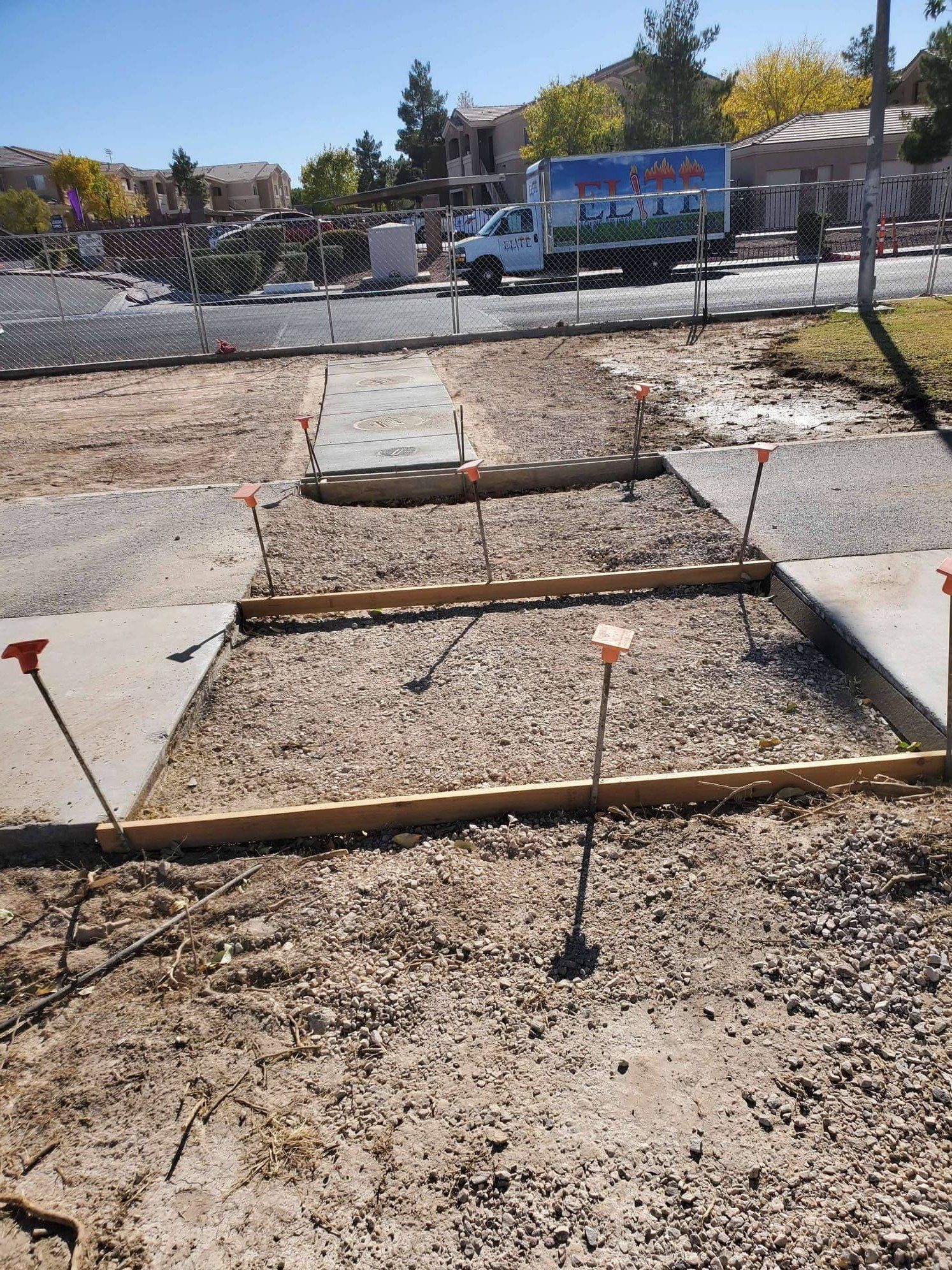 Construction site with wooden forms and gravel for a sidewalk. Stakes mark the boundaries.