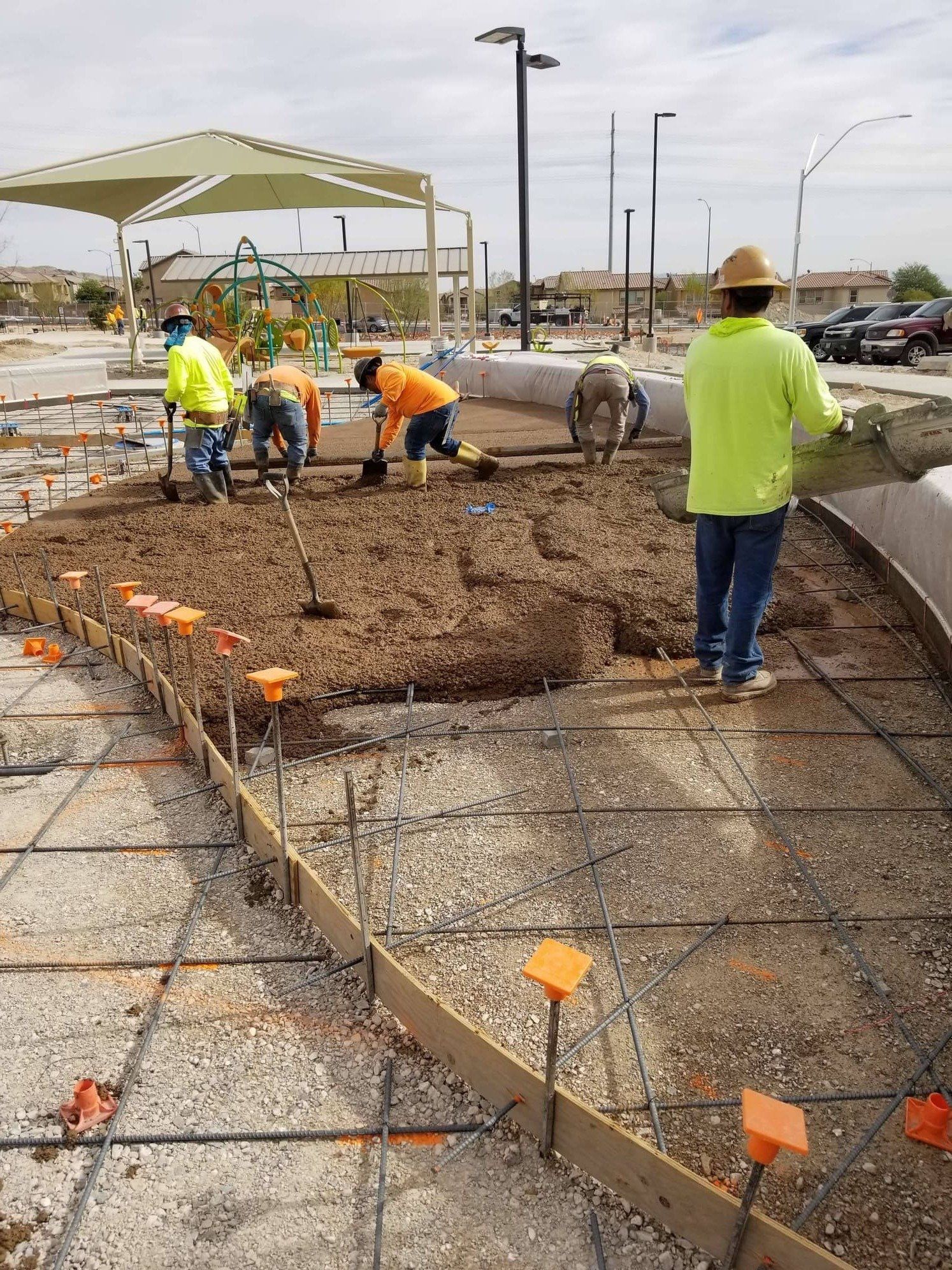 Construction workers pouring concrete at a park. They're using shovels and a wheelbarrow. Rebar and forms are visible.