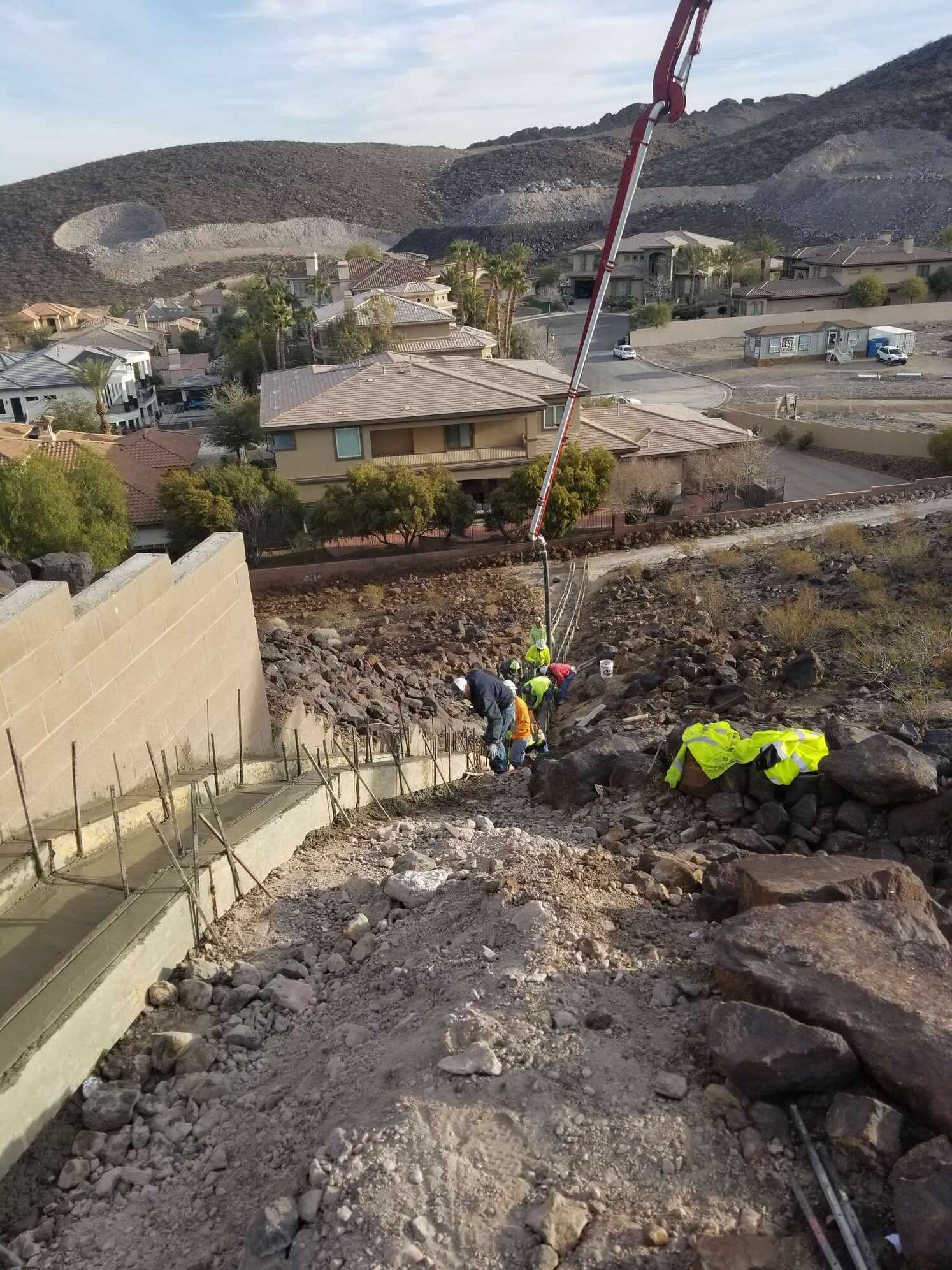 Construction workers pour concrete into a wall with a pump truck. Houses and mountains in the background.