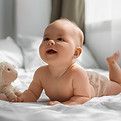Bébé allongé sur le ventre avec une peluche, regardant vers le ciel avec un sourire sur un lit blanc.