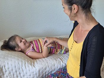Une femme caresse doucement un enfant allongé sur un lit, tous deux souriant dans une chambre lumineuse.
