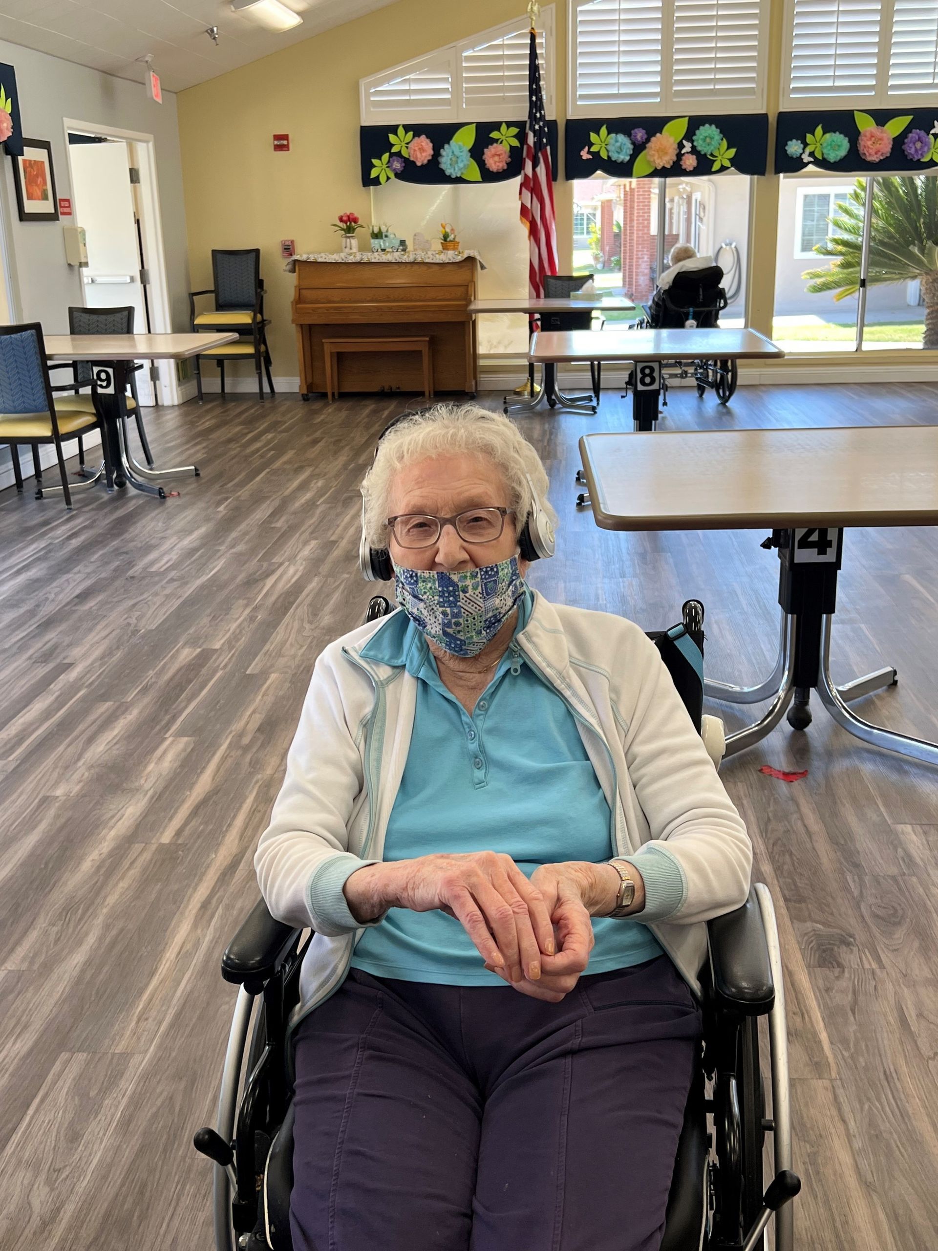 Elderly woman in a wheelchair wearing a mask, sitting in a room with tables, an American flag, and a piano.