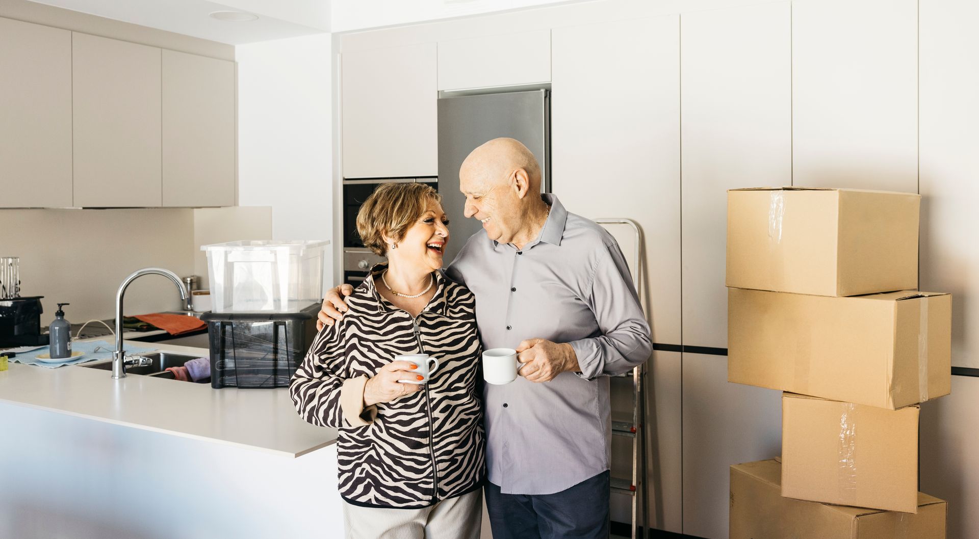 A happy senior couple enjoys coffee in their new kitchen, surrounded by moving boxes—downsizing neve