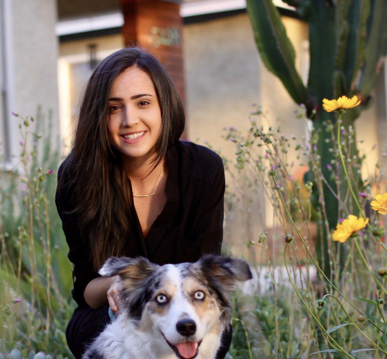 Woman with long dark hair smiling next to a blue-eyed dog in a garden, both looking at the camera.