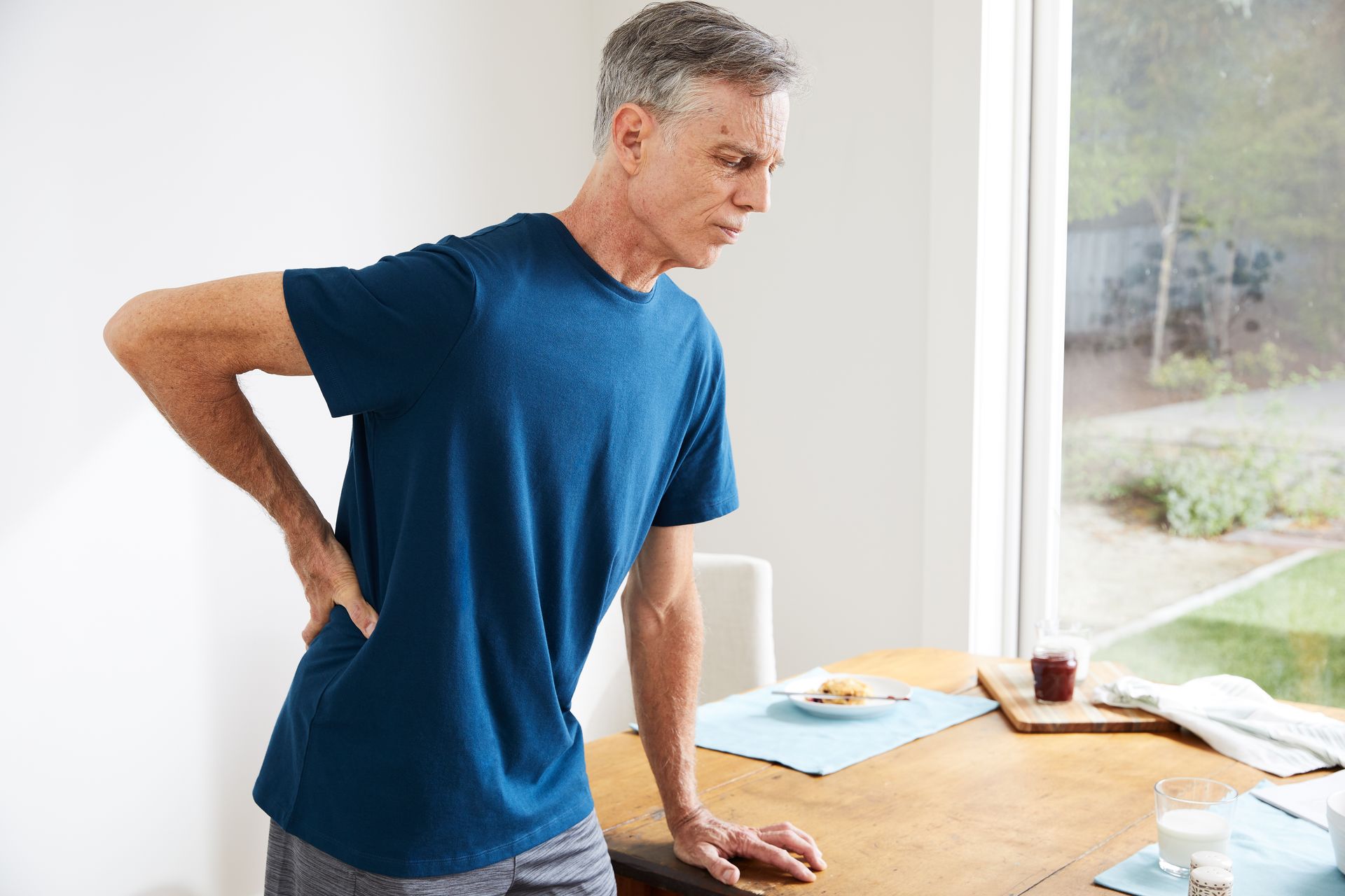 Man clutching his lower back, grimacing, near a table by a window.
