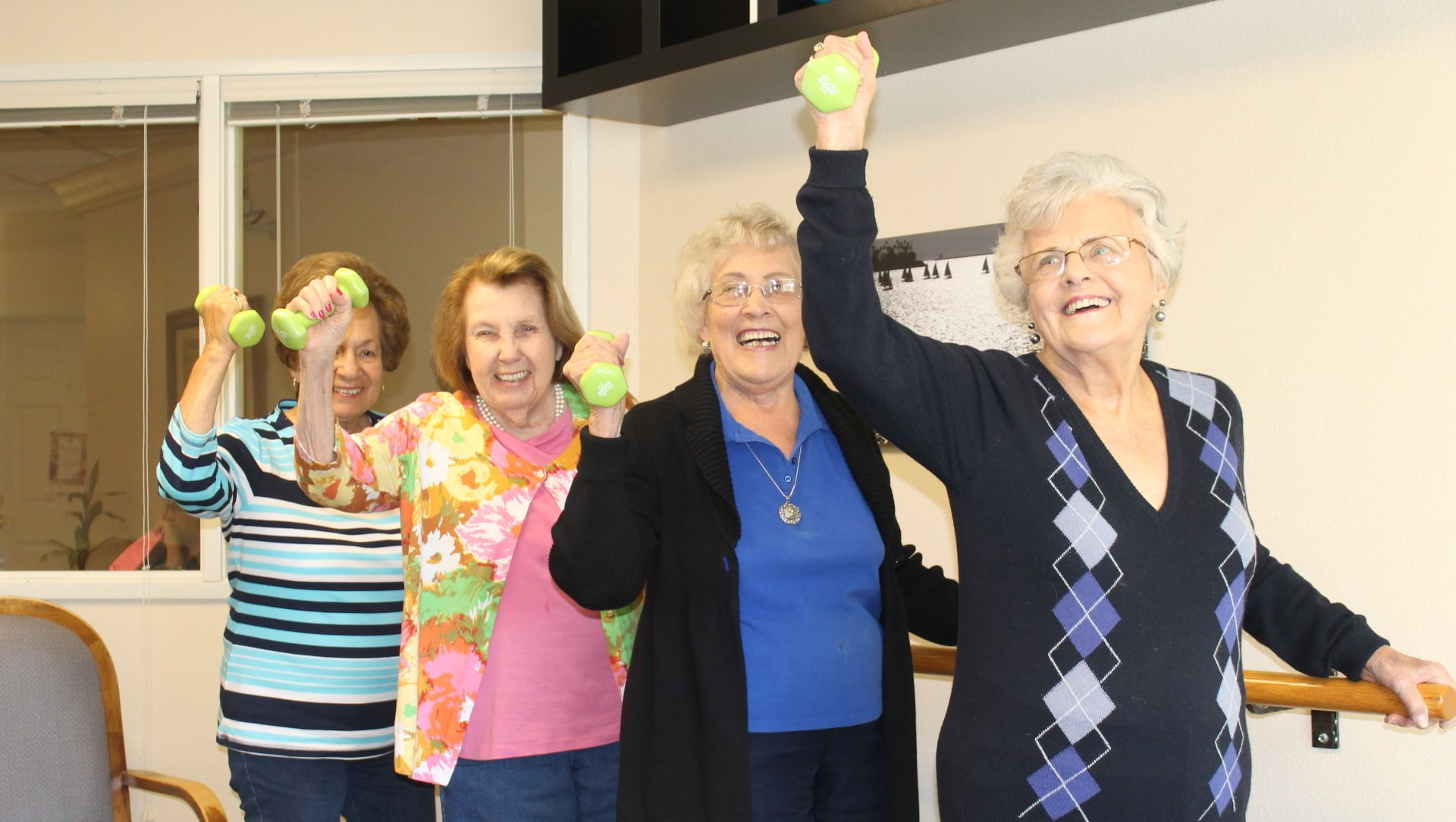 Four smiling elderly women lifting light green weights in a brightly lit room.