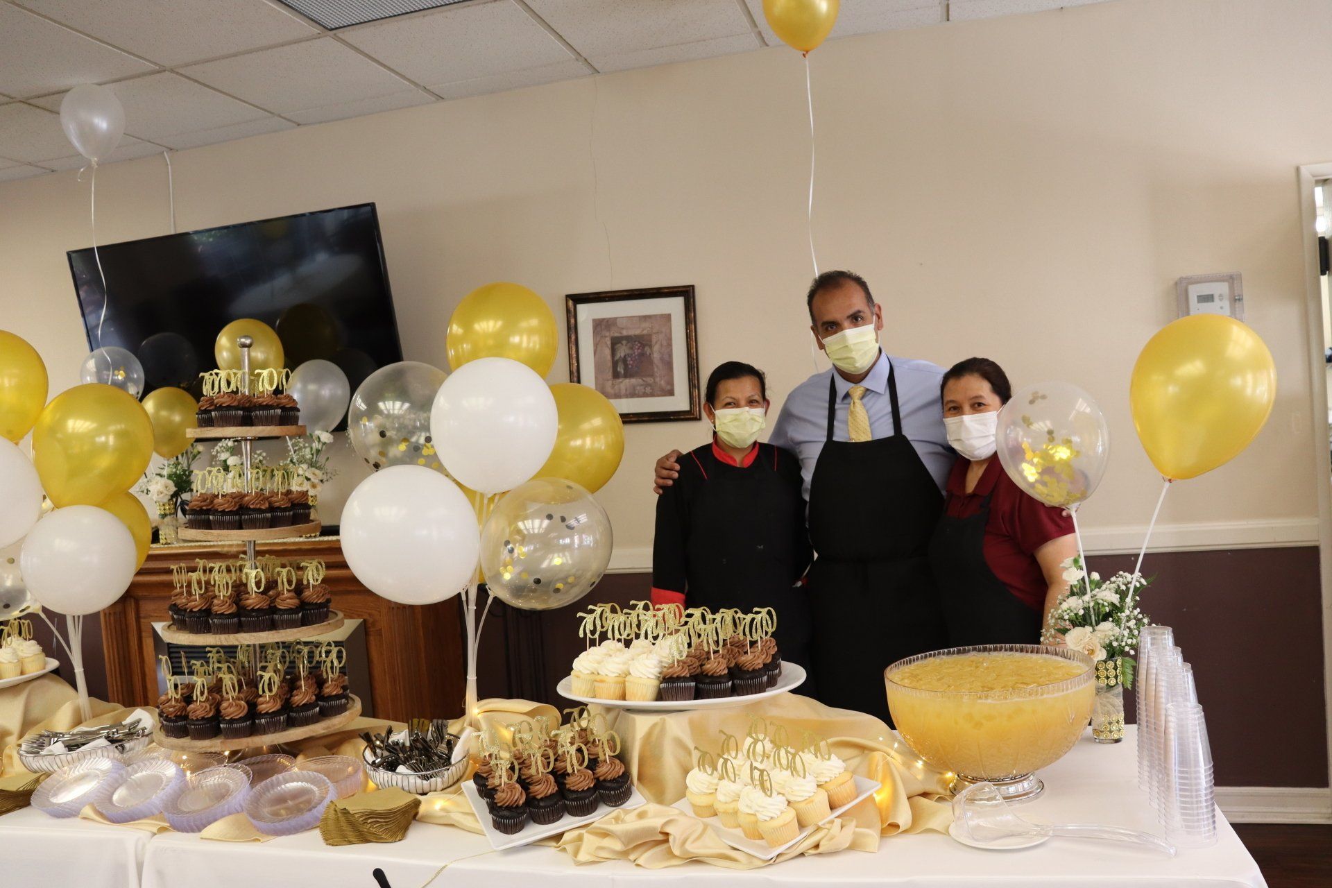Three masked people pose by a table of desserts with gold and white balloons.