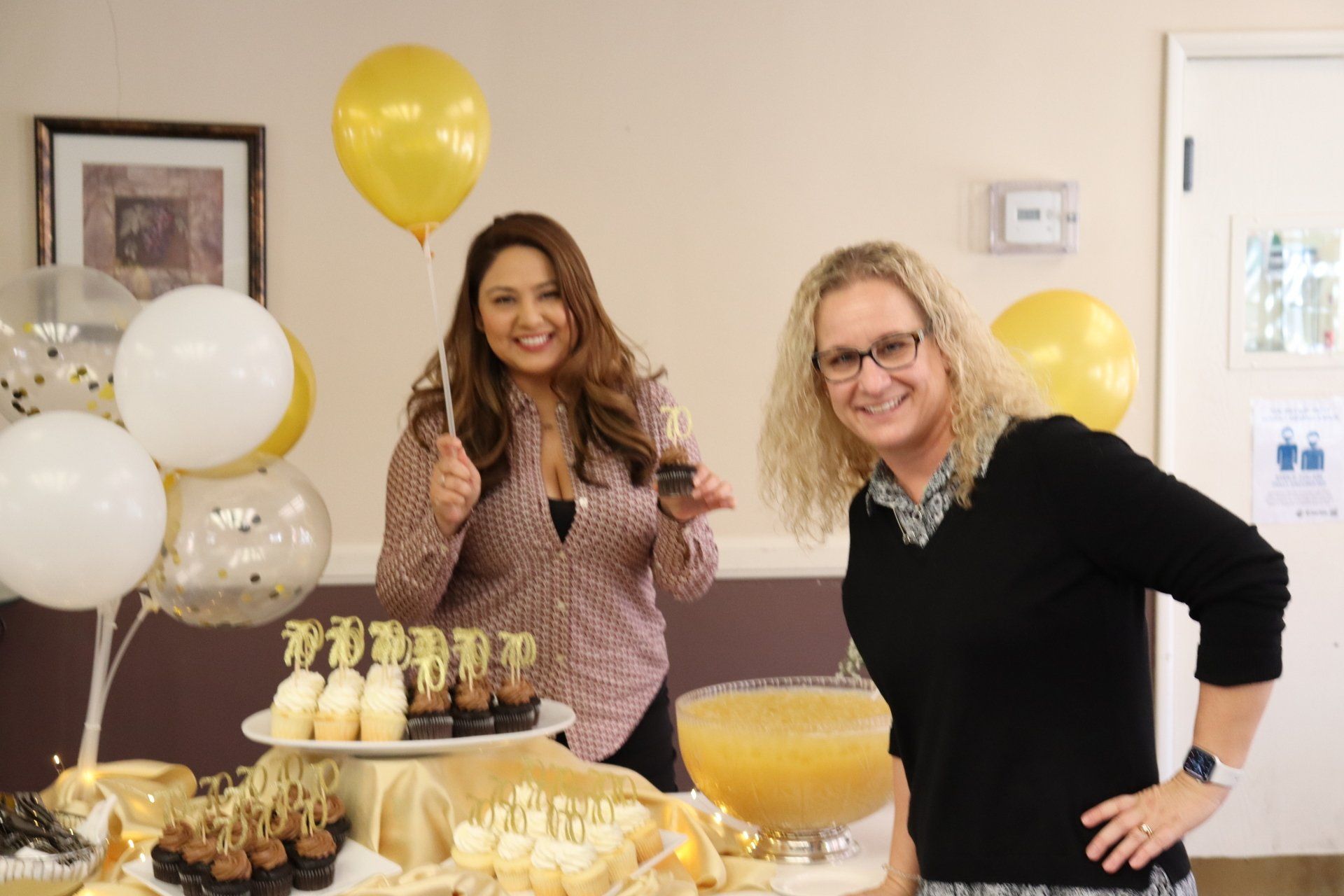 Two women smile at a party table with gold/white balloons and treats. One holds a cupcake and gives a thumbs up.