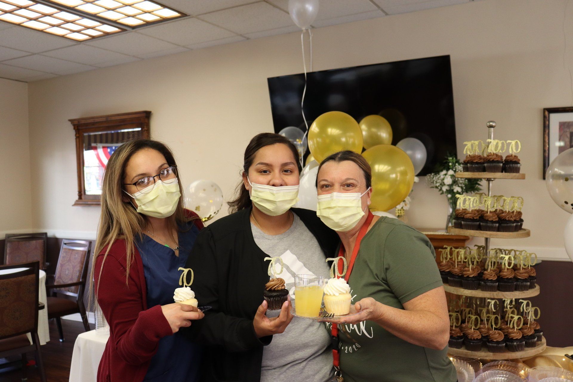 Three women wearing masks holding cupcakes, at a party with balloons and a tiered cupcake stand.