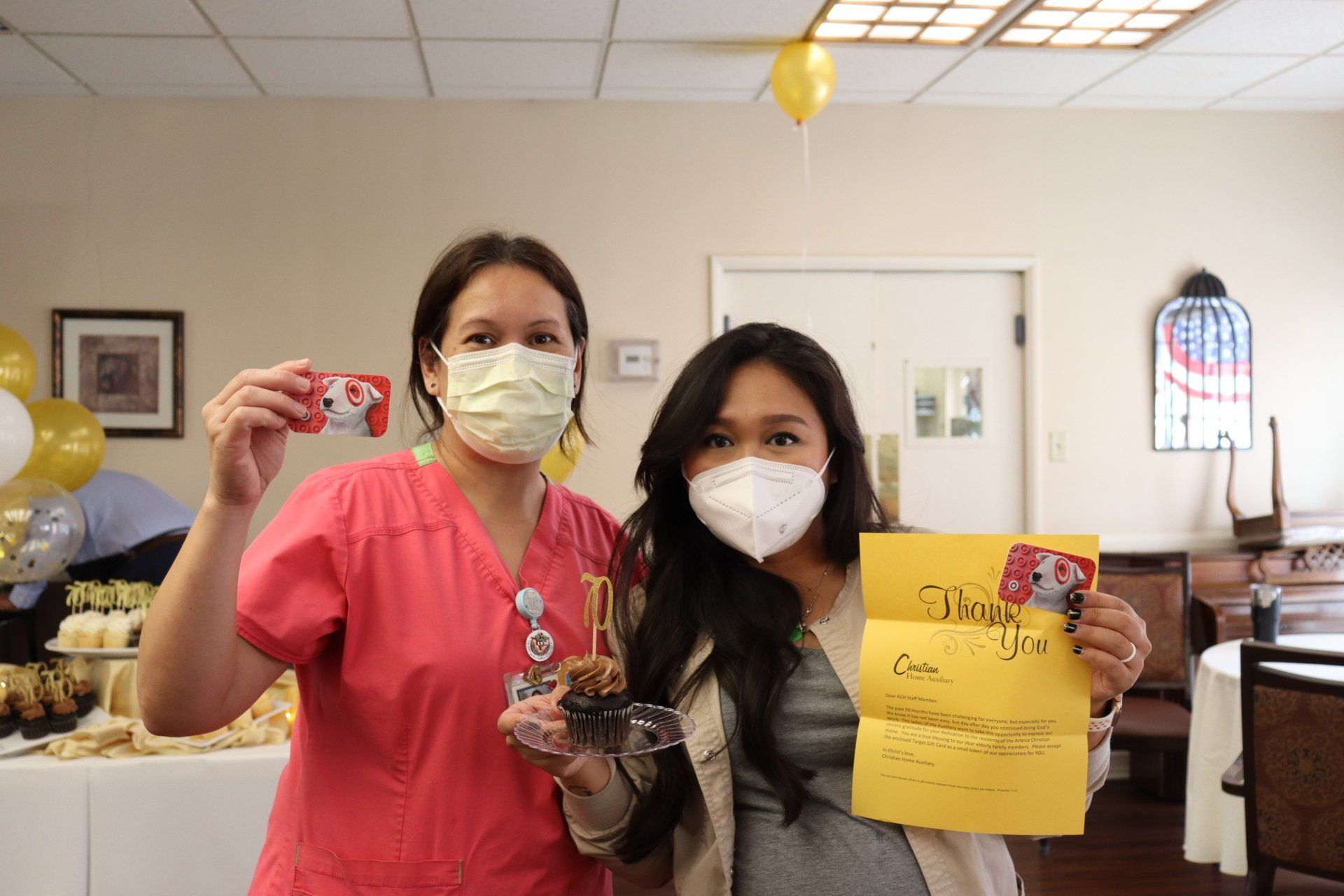 Two women in masks hold a gift card and thank-you note at a party. Balloons and treats are in background.