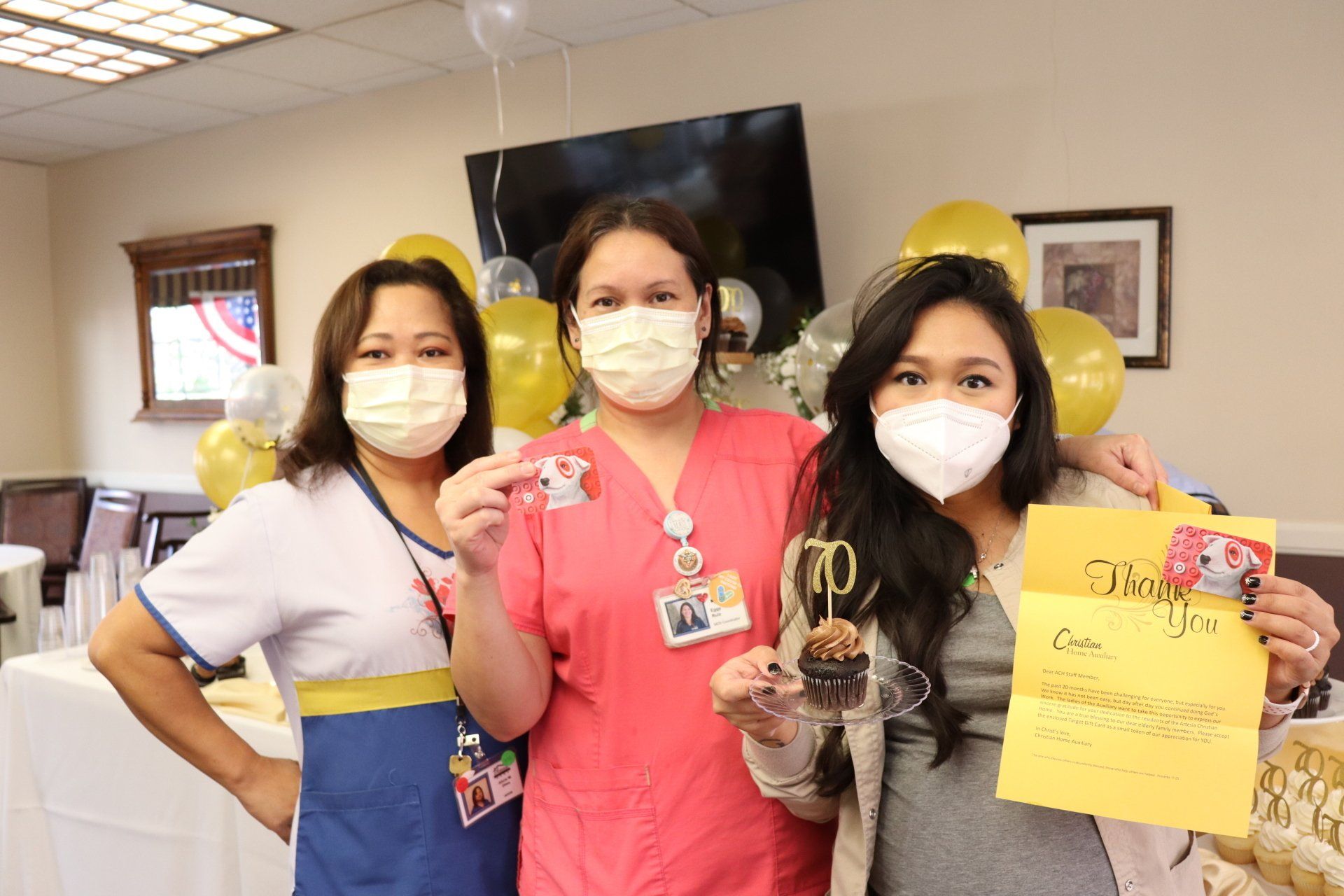 Three masked women celebrate a 70th birthday indoors. They hold a cupcake, a gift, and cards. Balloons in the background.