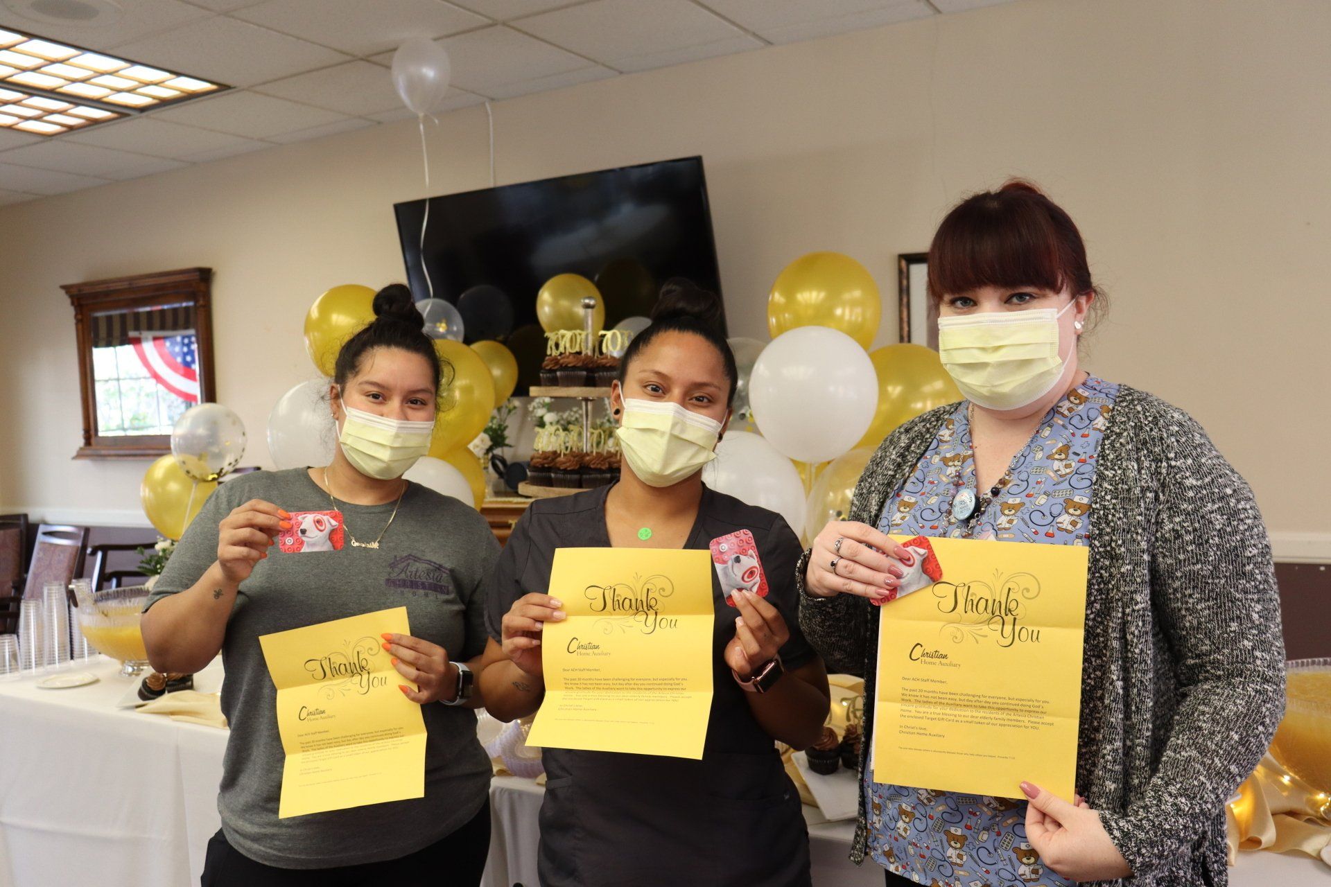 Three masked women hold cards and envelopes, smiling near balloons; celebratory setting.