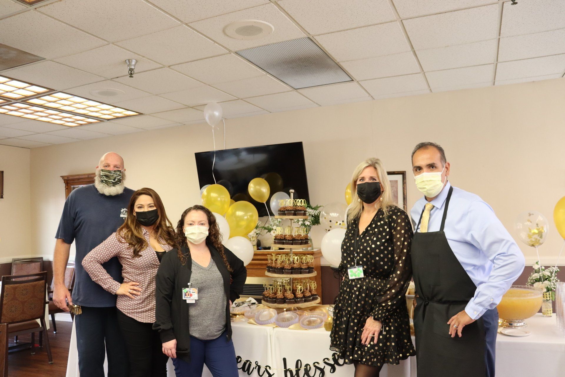 Group of people pose with a cake at an event; gold and white balloons, dressed up setting.