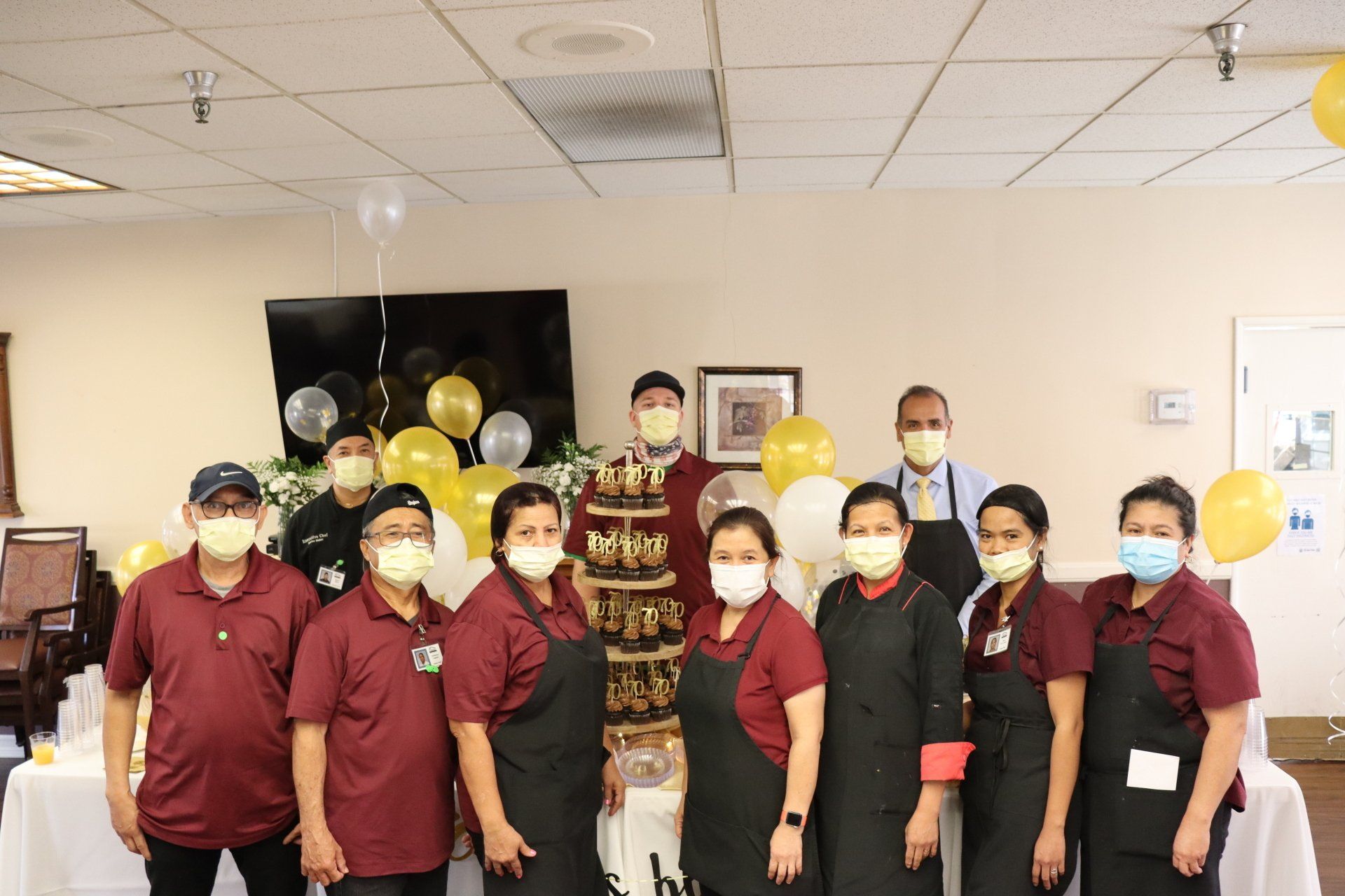 A group of people in maroon shirts and aprons pose with a cake, some wearing masks. Yellow balloons behind them.