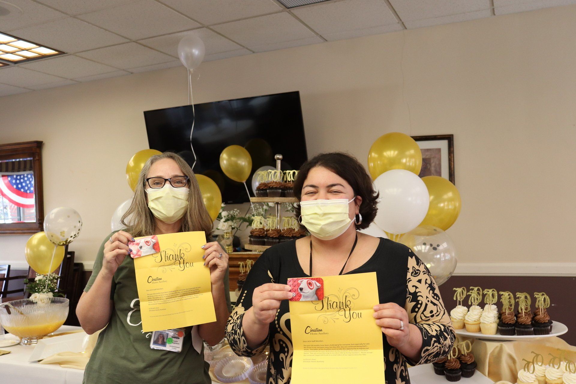 Two women in masks hold gift cards & cards, smiling. Decorated room with balloons & snacks.