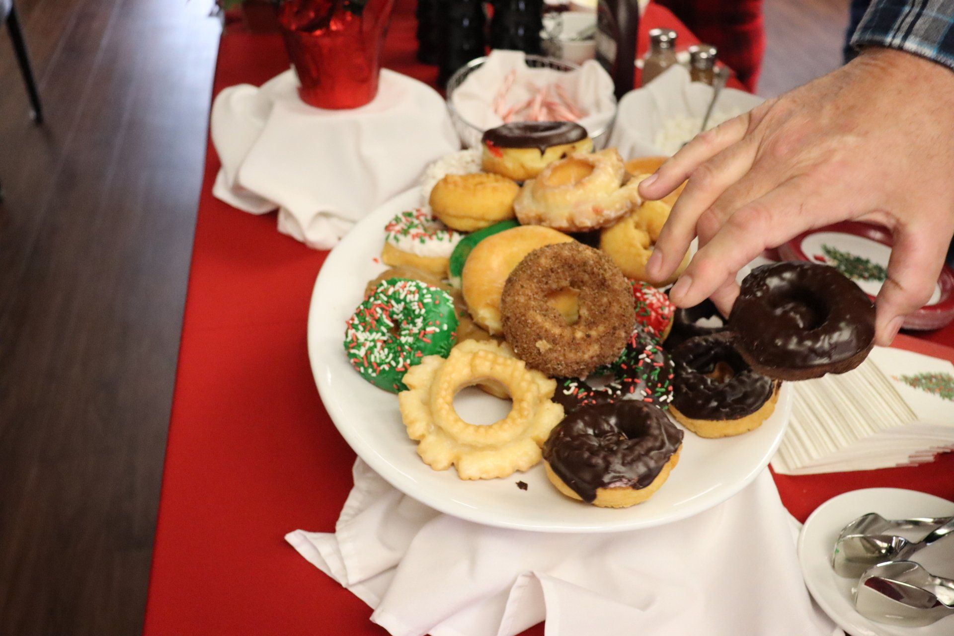 Person's hand reaching for a chocolate glazed donut on a plate piled high with assorted donuts on a red tablecloth.
