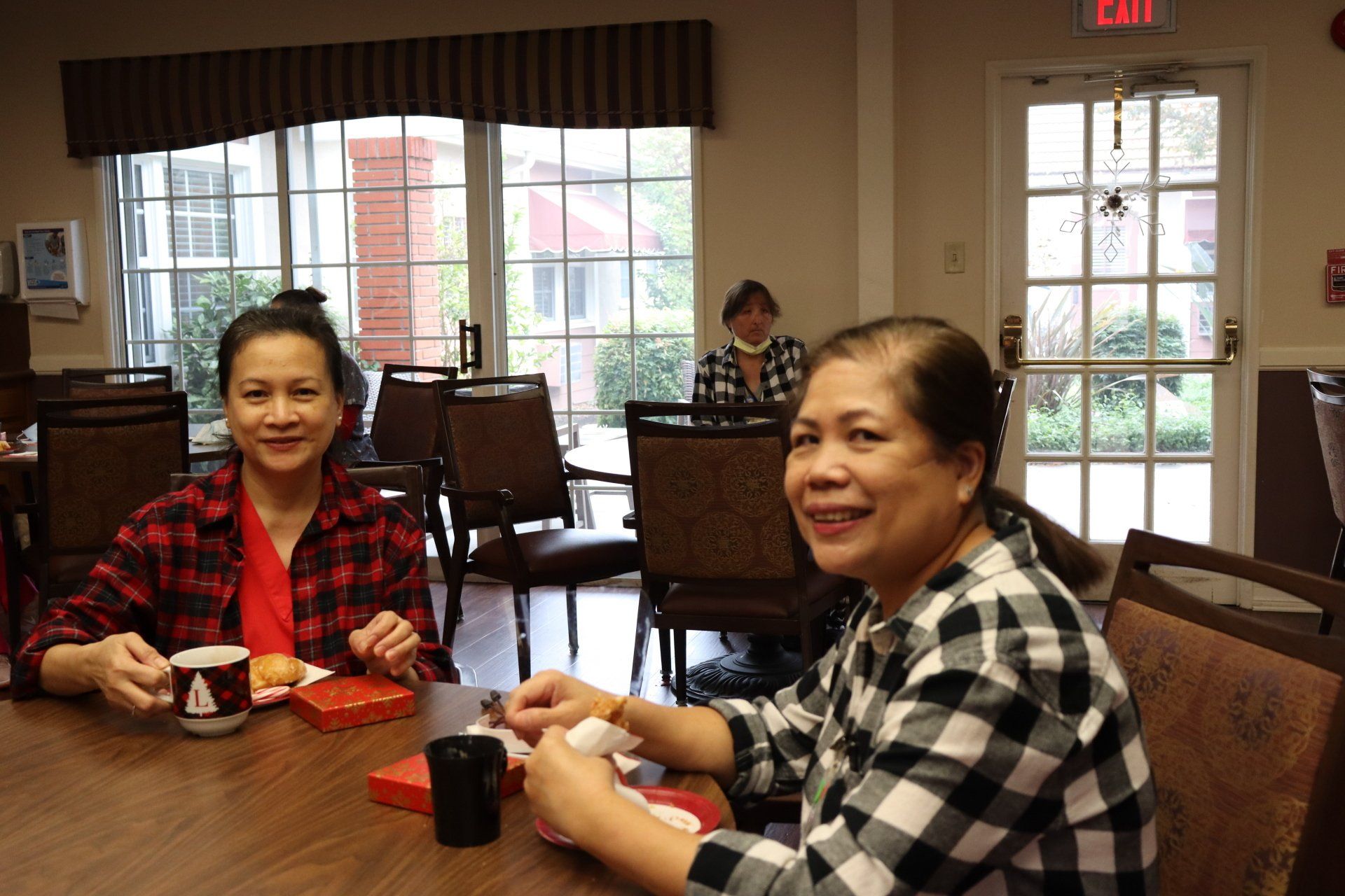 Two women in plaid shirts at a table, smiling and eating. Indoors, seated in a dining room with windows.